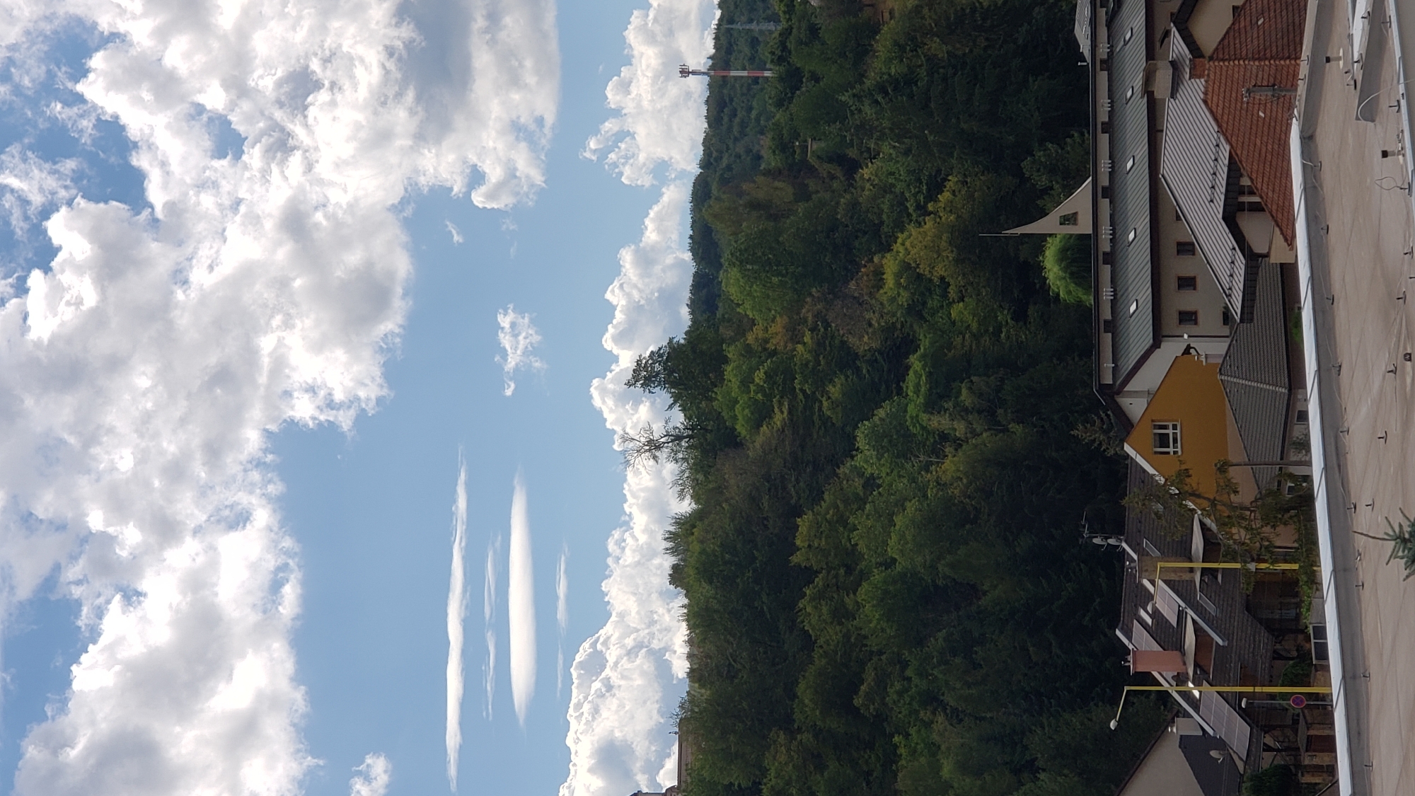 This is a vertical, outdoor, daytime photograph taken from a high vantage point overlooking a residential area and a densely forested hillside. The sky is a bright blue with scattered white cumulus clouds, some of which appear puffy and some as thin, horizontal layers. The top of the frame is dominated by the sky and clouds, with a prominent lenticular cloud formation visible in the upper left quadrant.
Below the sky, a dense green forest covers the hillside, with trees of varying shades of green, indicating different species or seasons. The foliage is thick and appears lush. A single, bare-branched tree stands out amongst the green canopy in the middle of the hillside. A red and white striped radio tower is visible on the far right of the hillside, and a faint power line tower can be seen to its left.
In the foreground, closer to the viewer, are the rooftops of houses and buildings. These structures are varied in design and color. Some have dark gray tiled roofs, while others have reddish-brown tiles. Several buildings feature solar panels on their roofs. A yellow building with a prominent white-framed window is visible, along with a tall yellow lamppost in front of it. The perspective suggests the photograph was taken from a higher building or elevated position looking down. The overall scene depicts a suburban or semi-rural environment bordering a natural landscape under a cloudy sky. There are no people visible in the frame.
