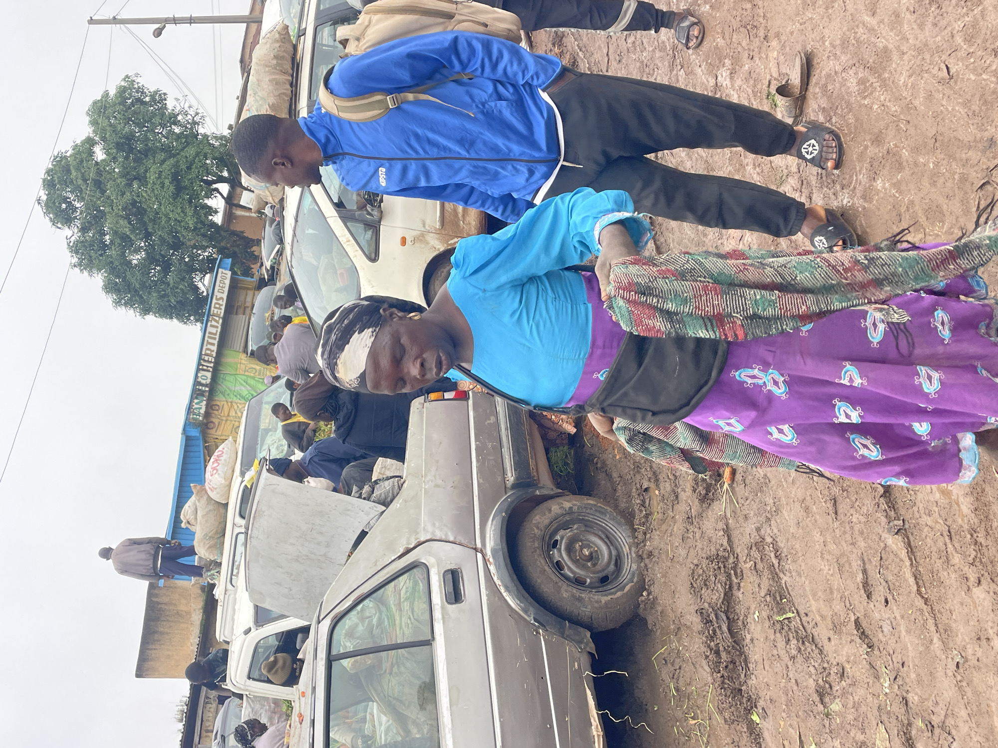 In Gura Top, Nigeria, on a muddy, overcast day, a marketplace scene unfolds around parked vehicles. A woman in a bright blue top and a purple skirt adorned with blue motifs walks towards the camera, draped in a colorful scarf. She wears a black fanny pack. Beside her, a young man in a blue athletic jacket and black pants with a backpack on looks to his right. Other individuals are visible in the background, some standing on top of vehicles laden with sacks of goods, and others near cars. A silver sedan with its hood open and trunk partially loaded is prominent on the left. The ground is uneven and heavily muddied, suggesting recent rainfall. Buildings with signage, including "FERTILIZERS DEPOT," and a large tree are visible in the background, indicating a commercial area. The overall atmosphere appears to be one of commerce and transport, likely related to agricultural produce given the sacks on the vehicles.