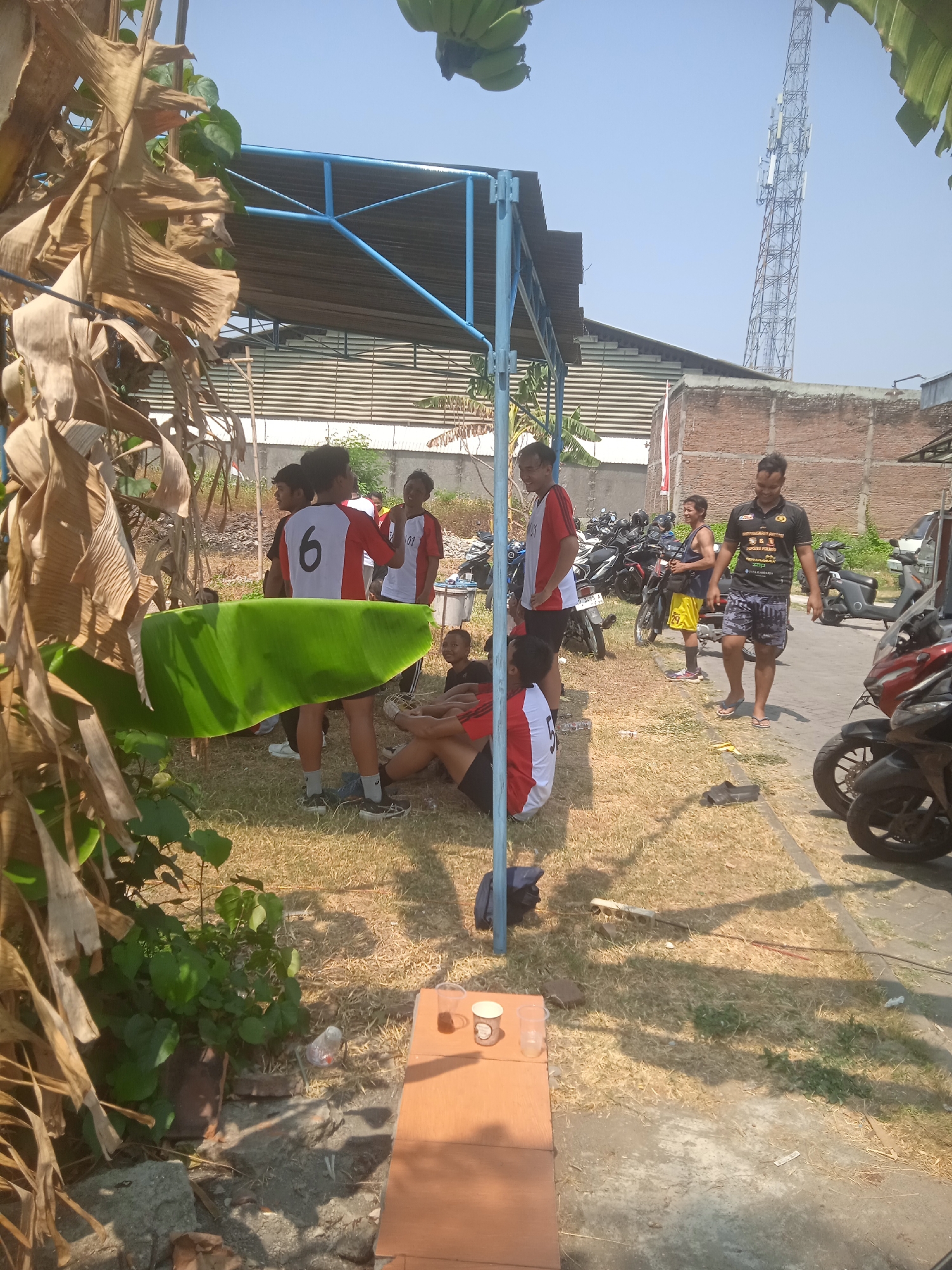 In Klodran, Indonesia, under a clear sky, a group of men are gathered outdoors near a covered structure with a blue frame and a corrugated metal roof. Several motorcycles are parked in the background. In the foreground, dry grass covers the ground, with a large, green banana leaf partially obscuring the view. One man, wearing a black t-shirt with text and a yellow logo, and patterned shorts, walks towards the right of the frame, wearing flip-flops. Another man in a yellow tank top and yellow shorts stands near a motorcycle. Several young men in athletic wear are also present. One is wearing a red and white shirt with the number '6' on the back. Another man is sitting on the ground, wearing a red and white shirt. A small wooden surface in the foreground holds a plastic cup with a dark liquid and an empty clear plastic cup. A cellular tower is visible in the distance.