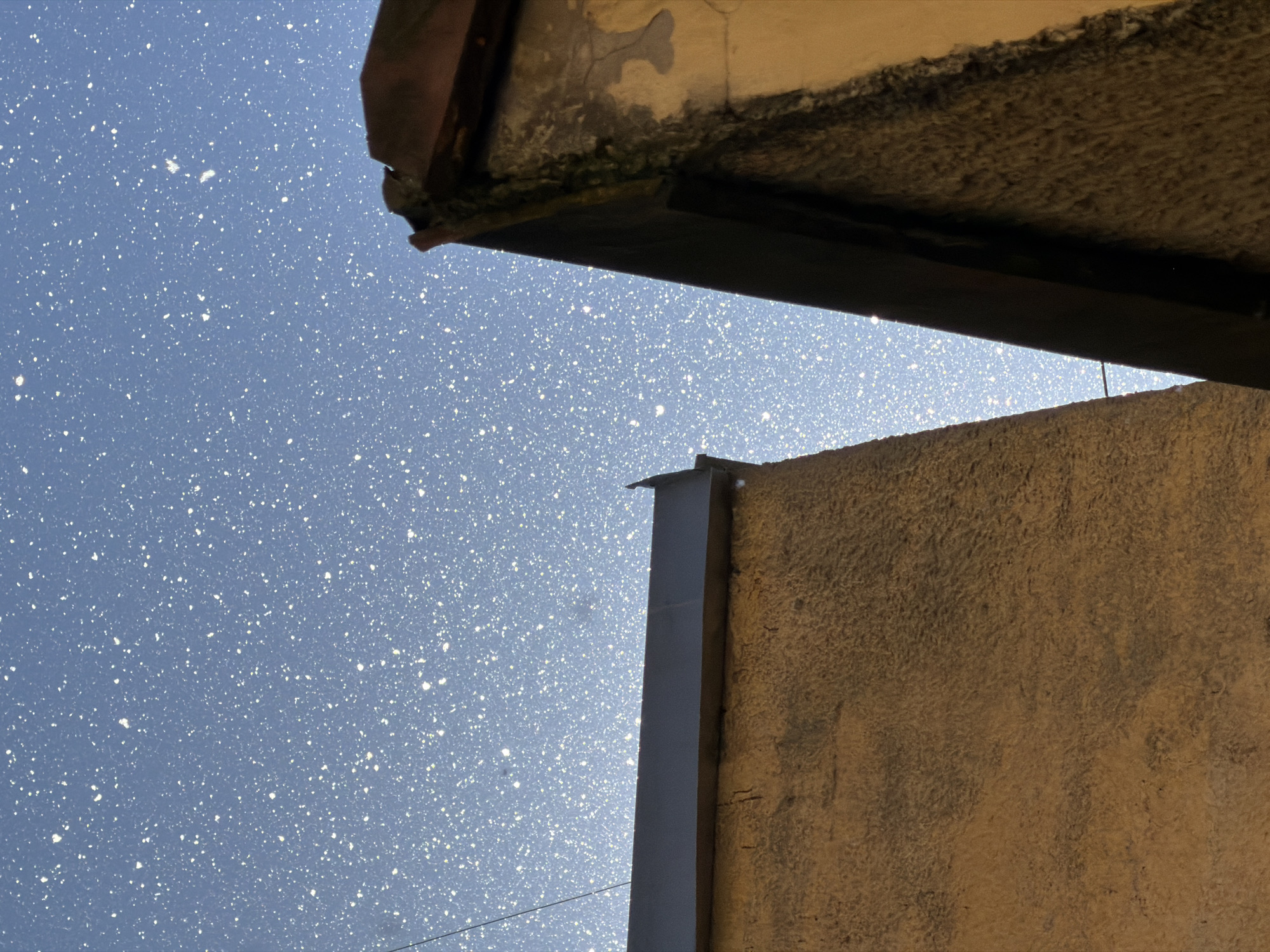 The image displays a low-angle view of two buildings against a blue sky filled with numerous small white dots, resembling stars or reflections. The building on the left is a rough-textured, light brown structure with a dark gray band along its top edge. The building on the right is a darker, more weathered structure with a reddish-brown tiled roofline. The sky is a gradient of light blue, with the white dots appearing more concentrated and brighter towards the top. A thin black line, possibly a wire, is visible in the upper left quadrant. The scene is likely captured at an elevated vantage point within the city of Saint Petersburg, Russia, given the architectural style of the buildings and the appearance of the sky. No people, actions, or interactions are depicted.