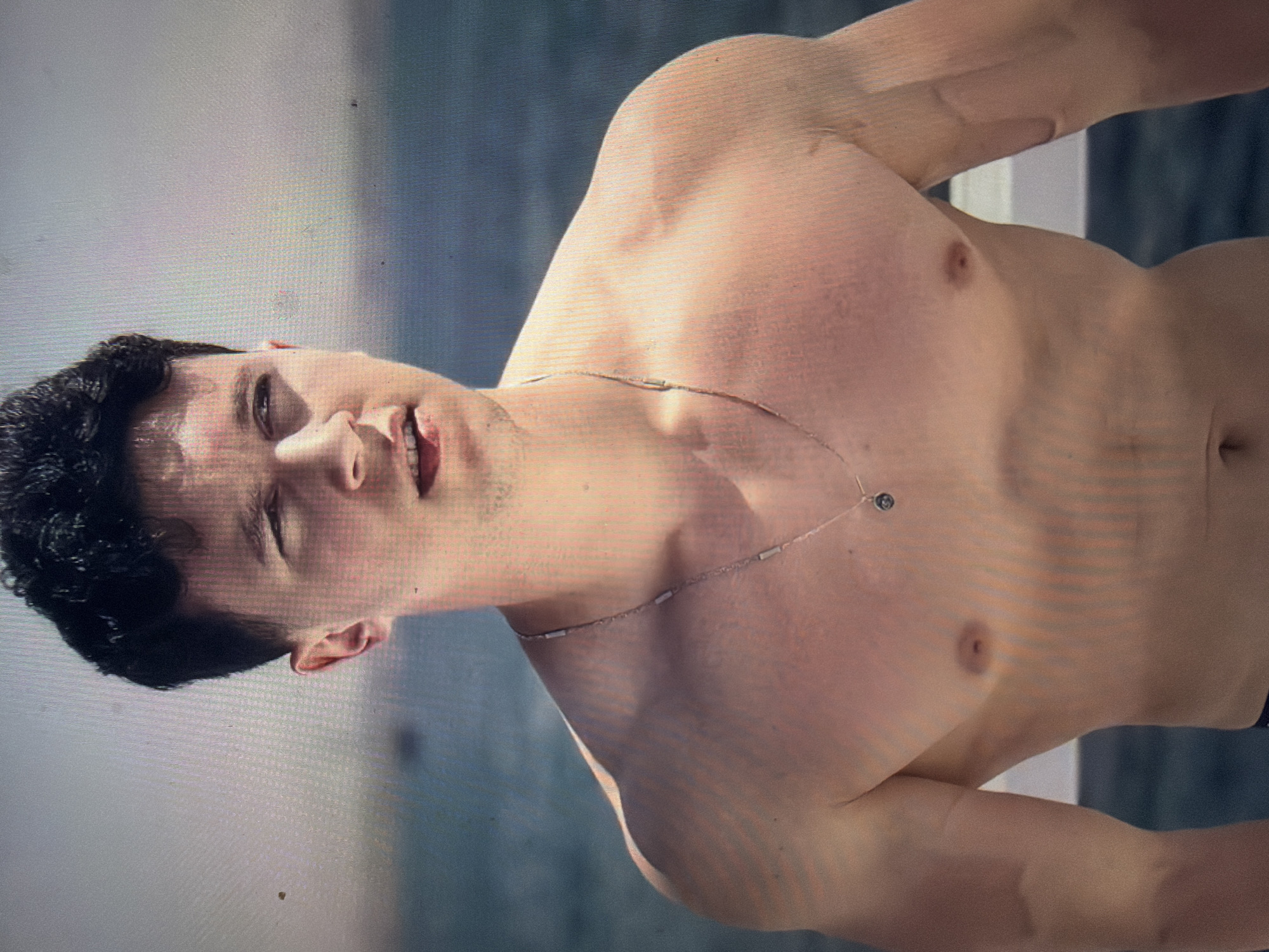 The image features a young adult male captured from the chest up, positioned outdoors against a blurred background of water and sky. The individual has short, dark, curly hair, a light skin tone, and a muscular build, particularly in the shoulders and chest. He is shirtless, wearing a thin, silver-toned chain necklace with a small, dark, circular pendant. His lips are slightly parted, revealing visible dental braces on his upper teeth, and his gaze is directed towards the viewer's right. Subtle facial hair, appearing as stubble, is present on his chin and upper lip. The background consists of a dark blue body of water in the lower portion, transitioning to a light gray sky above, indicating clear weather. A faint white structure, possibly a boat railing or deck, is partially visible behind him. The image exhibits a visible pixel grid pattern, suggesting it is a photograph of a screen or a highly compressed digital image. There are no identifiable visual markers within the image to confirm a specific location in the city of Shumen, Bulgaria.