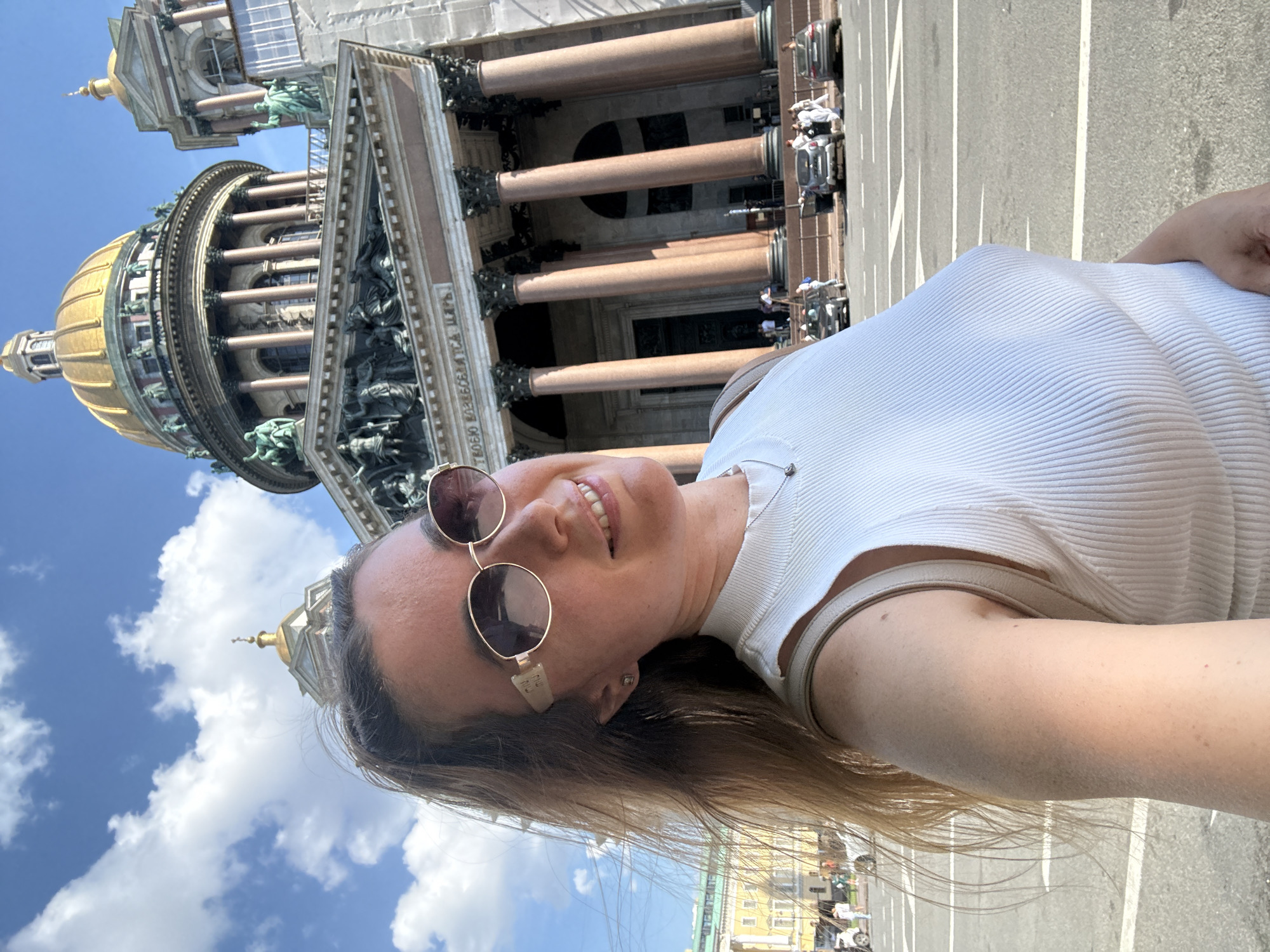 A selfie captures a young woman smiling brightly in front of a grand, domed cathedral on a sunny day. The woman, positioned in the foreground and appearing to hold the camera at a slight upward angle, has long brown hair and wears round-framed sunglasses. She is dressed in a white, ribbed top and displays a joyful expression with her teeth visible.
The magnificent building in the background features classical architecture with numerous large columns of a reddish-brown stone, topped with ornate green sculptural capitals. A prominent golden dome, characteristic of Saint Isaac's Cathedral in Saint Petersburg, Russia, dominates the upper left portion of the frame. Below the frieze of the cathedral, Cyrillic text is visible, reading "ТВОЕМУ ВЛАДЫЧЕСТВУ И ЦАРЮ" (To your Lordship and Tsar).
The sky is a clear blue with scattered white clouds, indicating pleasant weather. A paved street with white lines and faint glimpses of distant vehicles and people can be seen in the lower right background, grounding the scene in an urban setting. The image conveys a sense of happiness and leisure, likely a tourist enjoying the historic landmark.