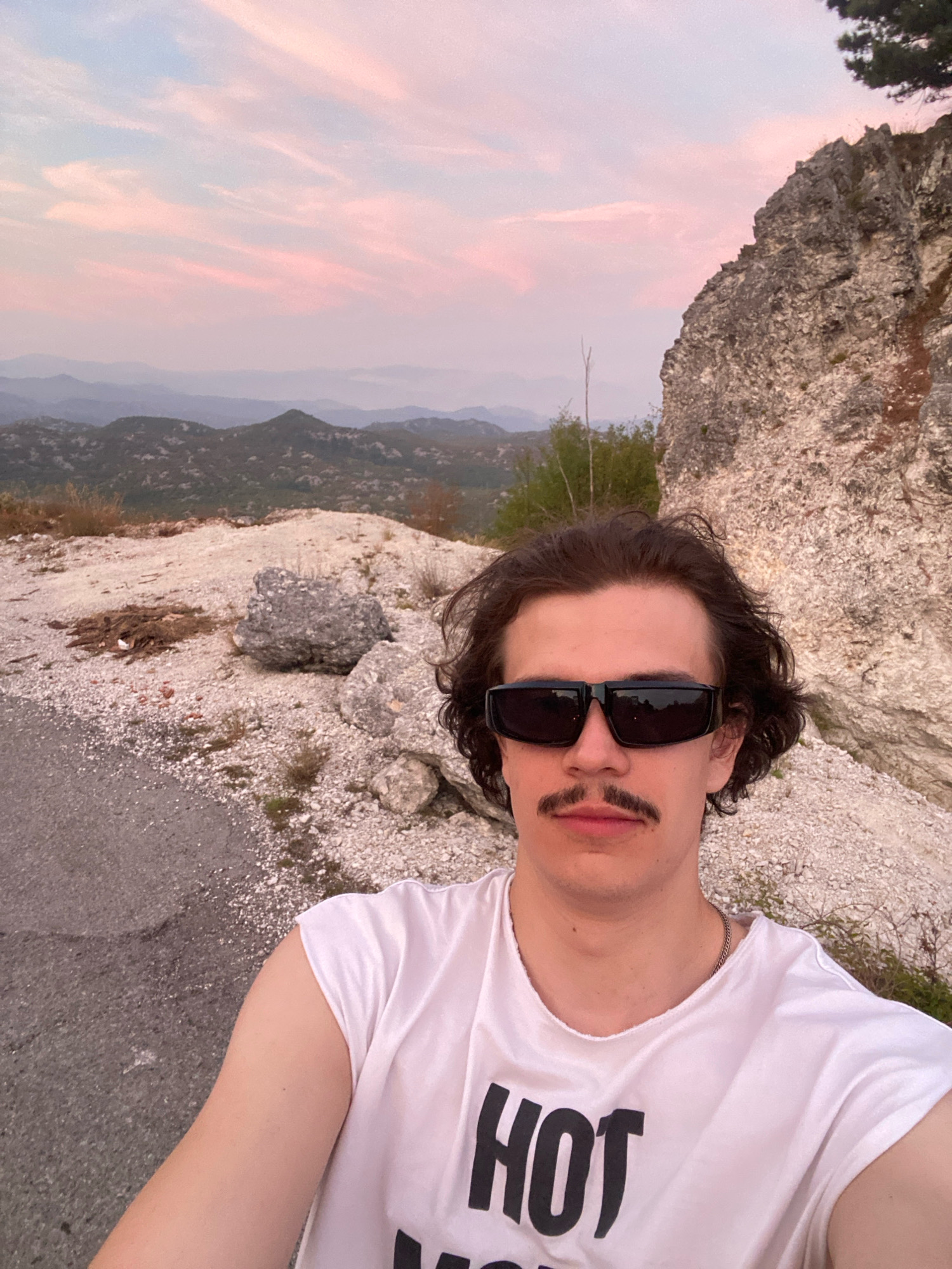 A young Caucasian male with dark, wavy hair, a prominent mustache, and black sunglasses poses for a selfie outdoors. He is wearing a white t-shirt with the word "HOT" in black block letters. He is standing on a gravelly roadside with a rocky cliff face to his right and a scenic view of rolling hills and mountains under a pink and blue sky in the background. The location is identified as Boguti, Montenegro.