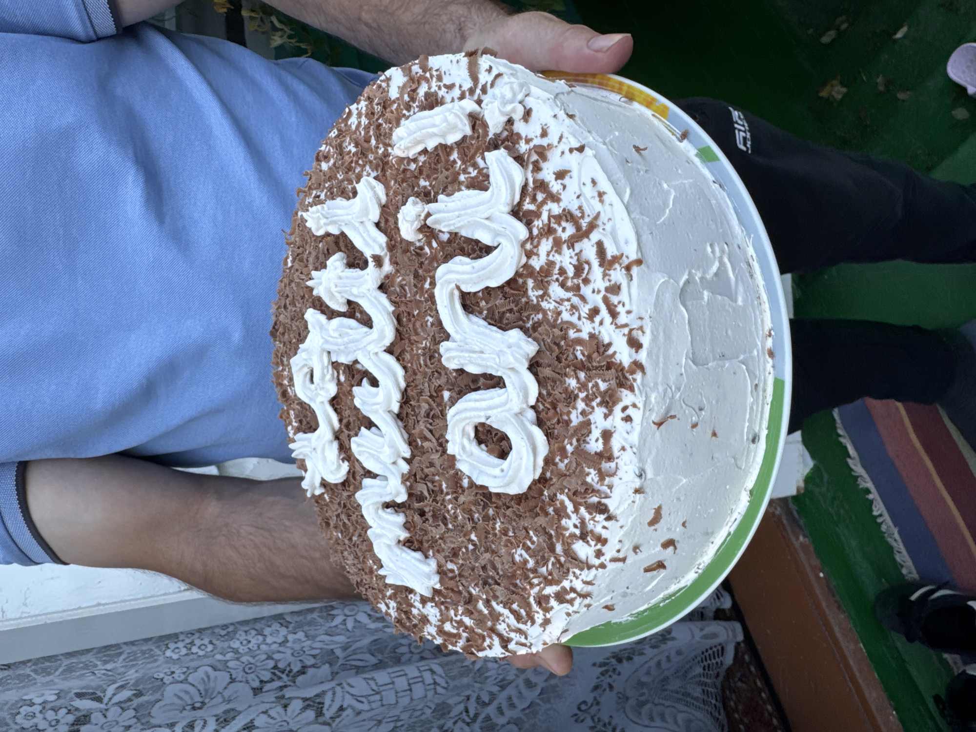 A person in a blue polo shirt is holding a round chocolate cake with white frosting and white icing lettering that reads "La mulți ani". The cake is decorated with chocolate shavings. The person is standing outdoors, with a lace curtain and a doorway visible in the background. The ground is covered with a colorful rug and green painted surface. The overall scene suggests a birthday celebration in Pănășești, Moldova.