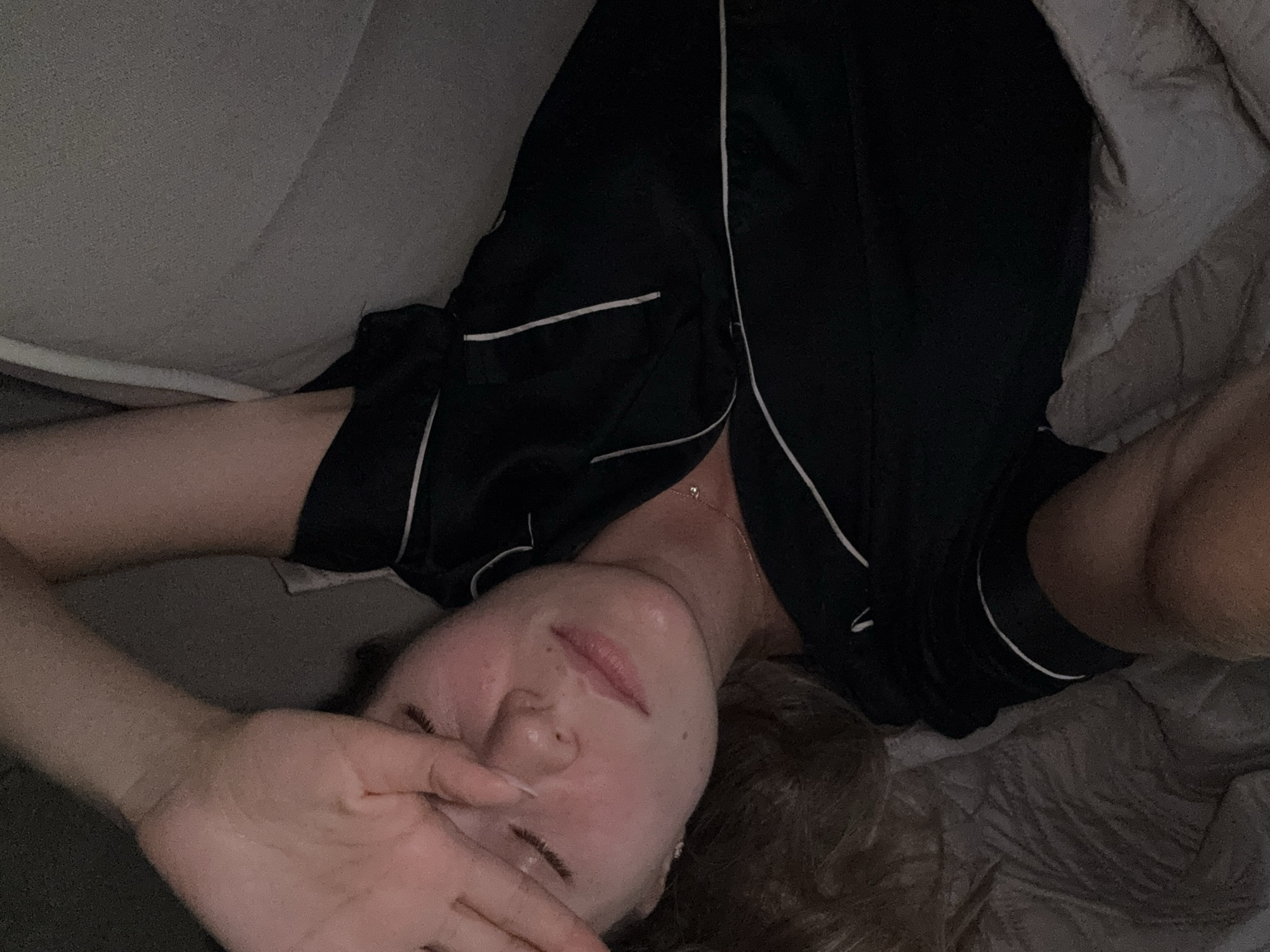 The image depicts a close-up, overhead shot of a young person lying down, their face partially obscured by their hand. They are wearing black pajamas with white piping. The lighting is dim, suggesting it might be nighttime or a poorly lit room. The person appears to be tired or sleepy, with their eyes closed and their hand resting on their face. The background consists of a textured blanket or duvet. There is no visible text in the image. The setting appears to be a bed or a comfortable resting place. The overall mood conveyed is one of relaxation or fatigue.