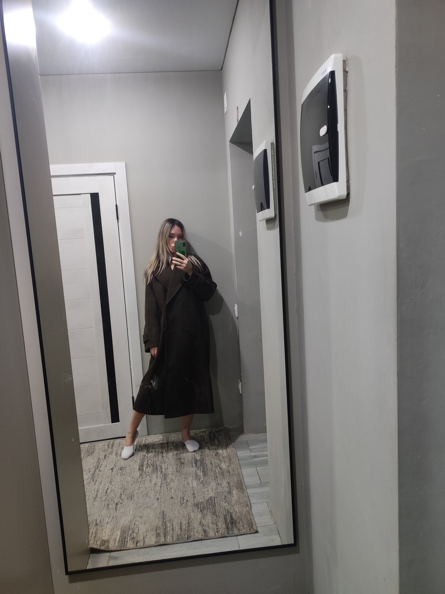 A woman with long blonde hair, wearing a long, dark brown coat and white slippers, is standing in front of a large mirror in what appears to be a hallway or entrance area. She is holding a green smartphone up to her face, seemingly taking a picture of herself in the mirror. The walls are painted in shades of gray. A white door with a black stripe is visible in the background. On the wall to the right of the mirror are two white and black plastic dispensers. A rug is seen on the floor. This scene is likely indoors but the specific location is not determinable. The city, Votkinsk, Russia, cannot be confirmed based solely on the image.