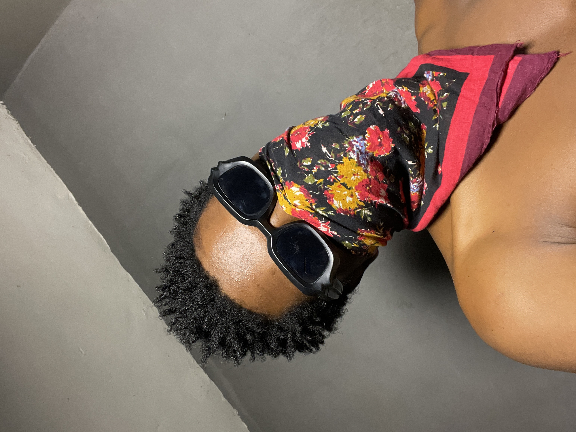 A close-up, waist-up shot shows a person wearing dark, angular sunglasses and a floral bandana over their face. The bandana has a black background with red, yellow, and blue floral patterns, bordered by red and burgundy edges. The person has dark, short, curly hair and visible skin on their shoulders and upper chest. The background is a plain, grey wall with a diagonal, lighter grey line running across the top left, suggesting a corner or a beam. The lighting is dim, casting shadows and highlighting the texture of the skin and bandana. The scene appears to be indoors, in a dimly lit room, potentially a residential setting in Nkpor, Nigeria.