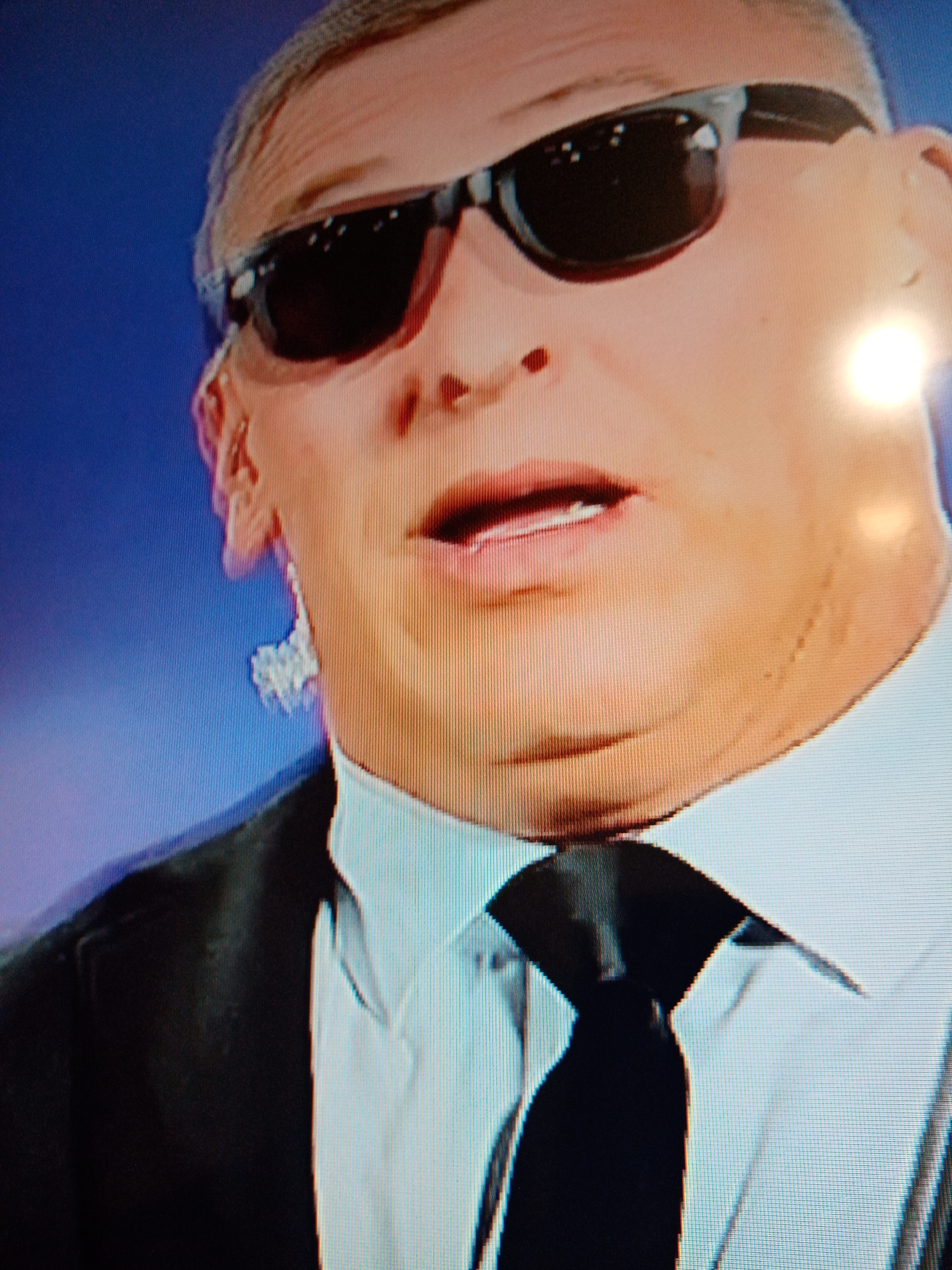 A close-up shot shows a man wearing sunglasses, a white shirt, a black tie, and a black suit jacket. He also has what appears to be a clear earpiece in his left ear. The background is a solid blue. The image appears to be a still from a video or television screen. There is no indication of the location being Elektrostal, Russia.
