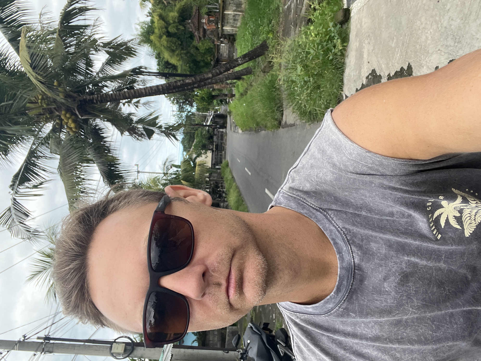 A selfie-style photograph shows a man wearing sunglasses and a gray t-shirt with a palm tree graphic. He is positioned with a street and tropical foliage in the background, including palm trees. The location is likely Sukawati, Indonesia, as indicated by the tropical scenery and potentially Balinese architectural details partially visible in the background. A paved road stretches into the distance, flanked by greenery. Utility poles are present alongside the road. A motorcycle or scooter is partially visible at the bottom of the frame.