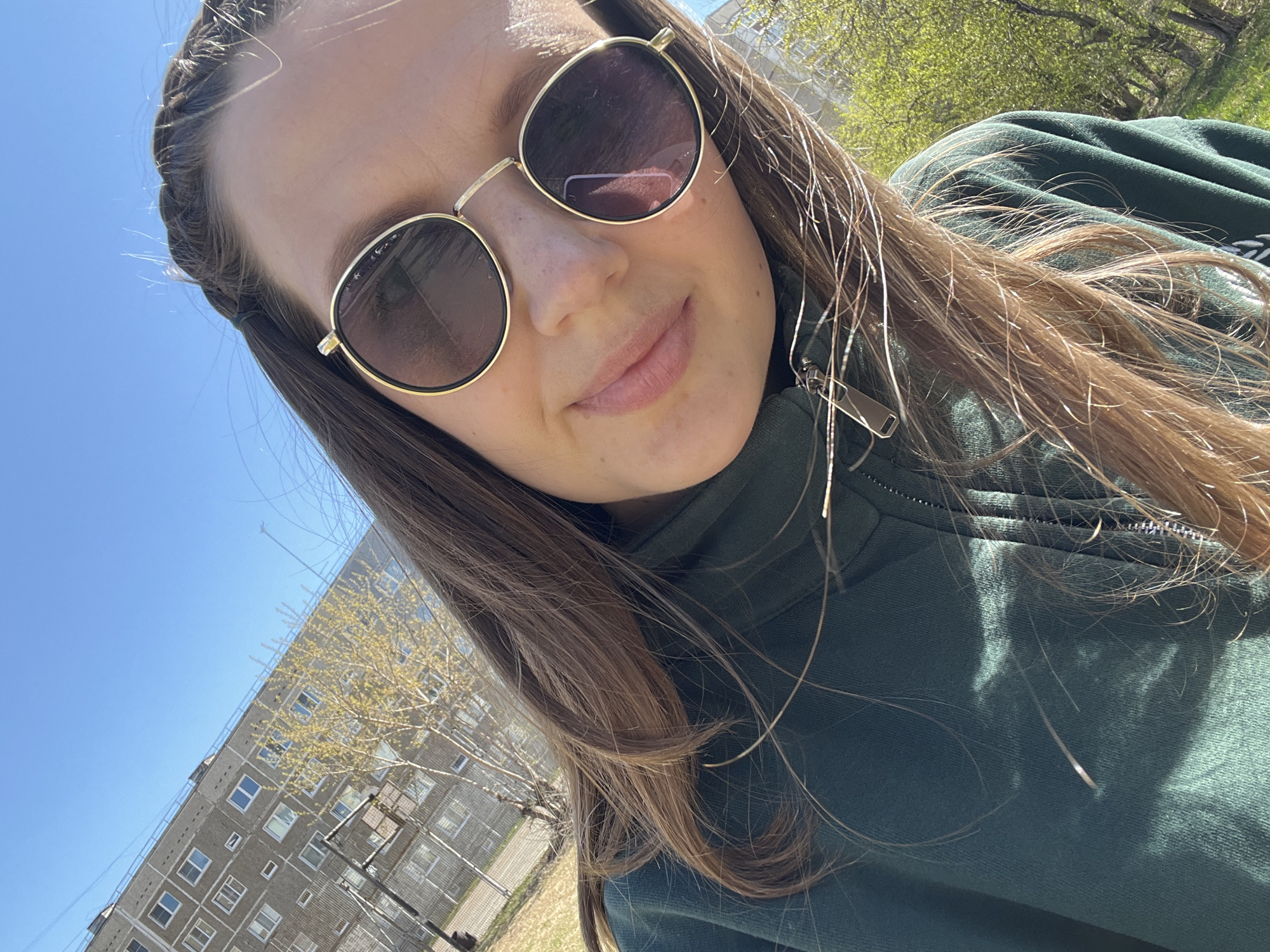 A selfie of a woman wearing sunglasses and a green zip-up sweatshirt, taken outdoors in Chaikovsky, Russia. Her hair is partially braided. In the background, there is a large, Soviet-era apartment building and bare trees against a blue sky. The image is slightly tilted.