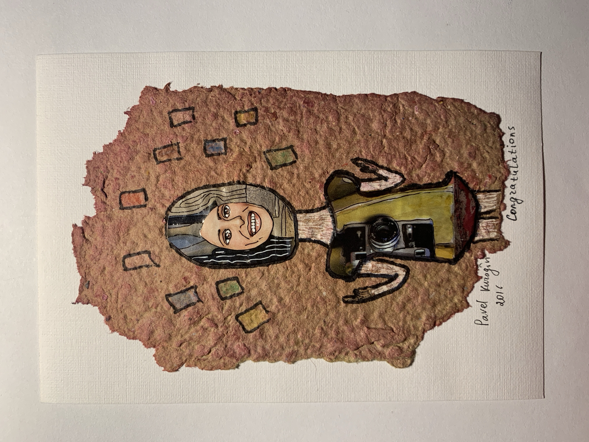 The image is a mixed-media artwork on textured white paper. The central subject is a stylized, collage-like figure with a torn, reddish-brown paper background that resembles a cloud or a messy explosion. This background is decorated with several small, rectangular shapes in muted colors, outlined in black.
The figure has a round face collaged from a photograph of a smiling woman. The rest of the head is drawn with black and white stripes within a helmet-like shape. The neck is a thin strip of textured paper. The body is depicted with a yellow dress that has puffed sleeves and a red hemline. On the chest of the figure is a detailed illustration of a vintage camera. The arms are outstretched, with hands drawn with simple lines, one index finger pointing upwards.
Below the artwork, handwritten text in black ink reads "Pavel Kurogin", "2016", and "Congratulations". The text is placed in the lower part of the white paper.
There is no identifiable location in the city of Sergeevo, Russia, depicted or suggested by the artwork. The image is a studio or indoor photograph of the artwork itself, with the textured white paper serving as a neutral backdrop.