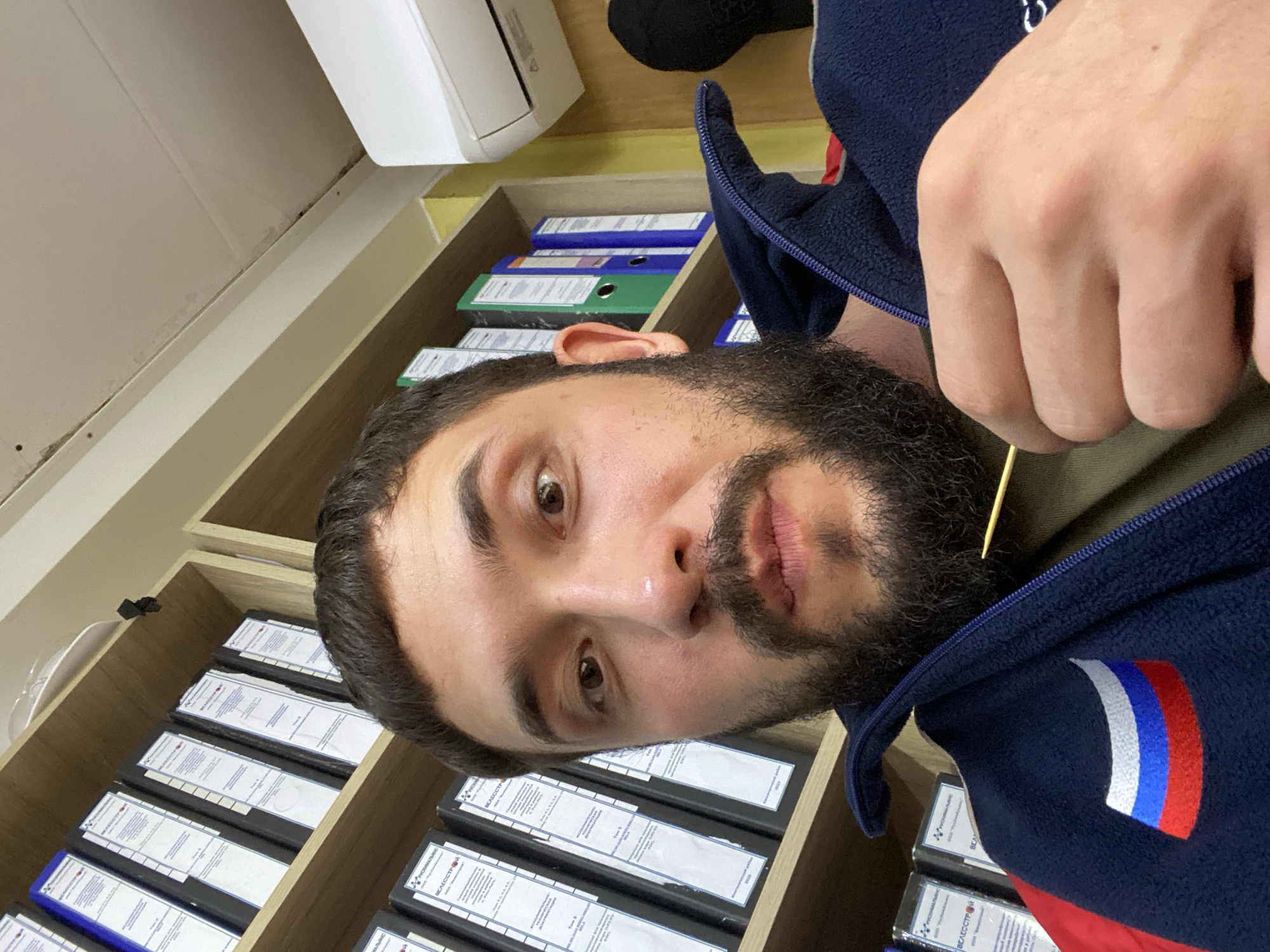 A man is taking a selfie in an office environment. He has dark hair, a beard, and is wearing a dark blue fleece jacket with a Russian flag patch on the left chest. He is holding a thin, light-colored object, possibly a toothpick, near his mouth. In the background, there are shelves filled with binders and an air conditioning unit mounted on the wall. The office appears to be located in Luzhitsy, Russia.