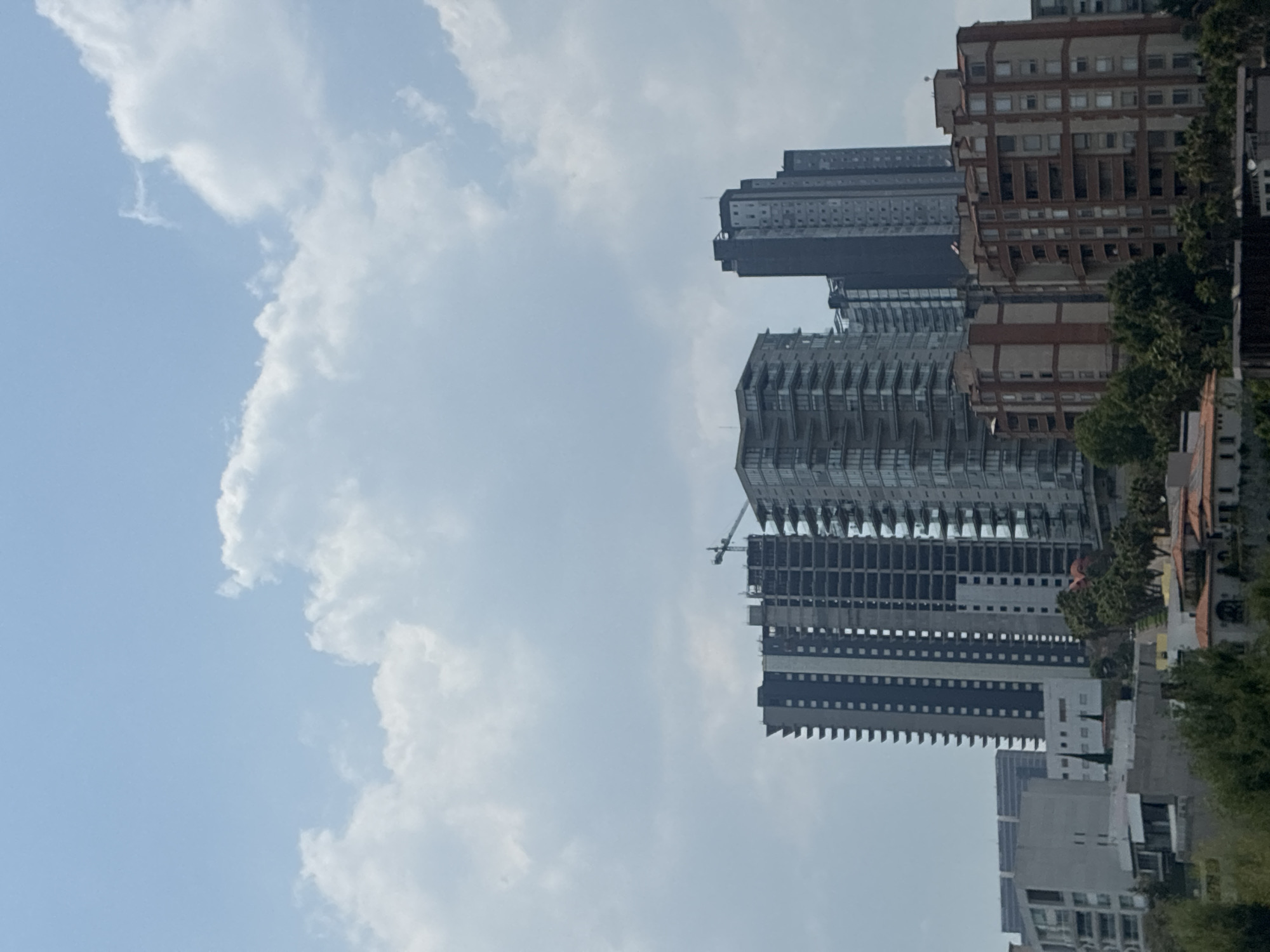 The image shows a partly cloudy sky above a mixed urban landscape, likely located in Huixquilucan de Degollado, Mexico. Tall buildings of varying architectural styles dominate the skyline. Some buildings appear to be modern high-rises, while others have a more traditional design. At least one building is under construction, with a crane visible. The lower foreground is occupied by trees and shrubs, indicating the presence of green spaces within the urban environment.