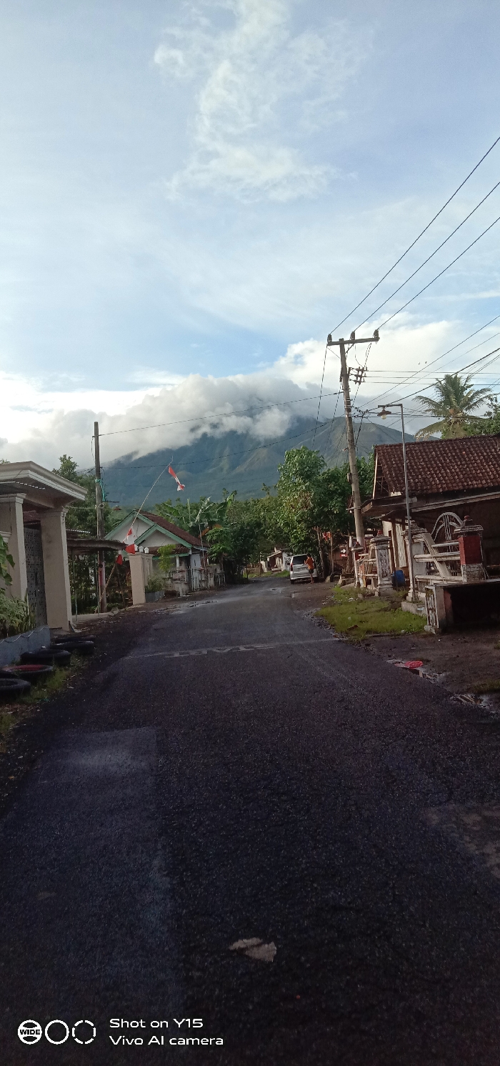 A street runs through a rural area with houses on both sides. A large mountain looms in the background, its peak shrouded in clouds. The sky above is partly cloudy. Indonesian flags are visible near some of the houses. The street is paved with asphalt. A power line runs along the right side of the street. The camera watermark is present on the bottom of the image.