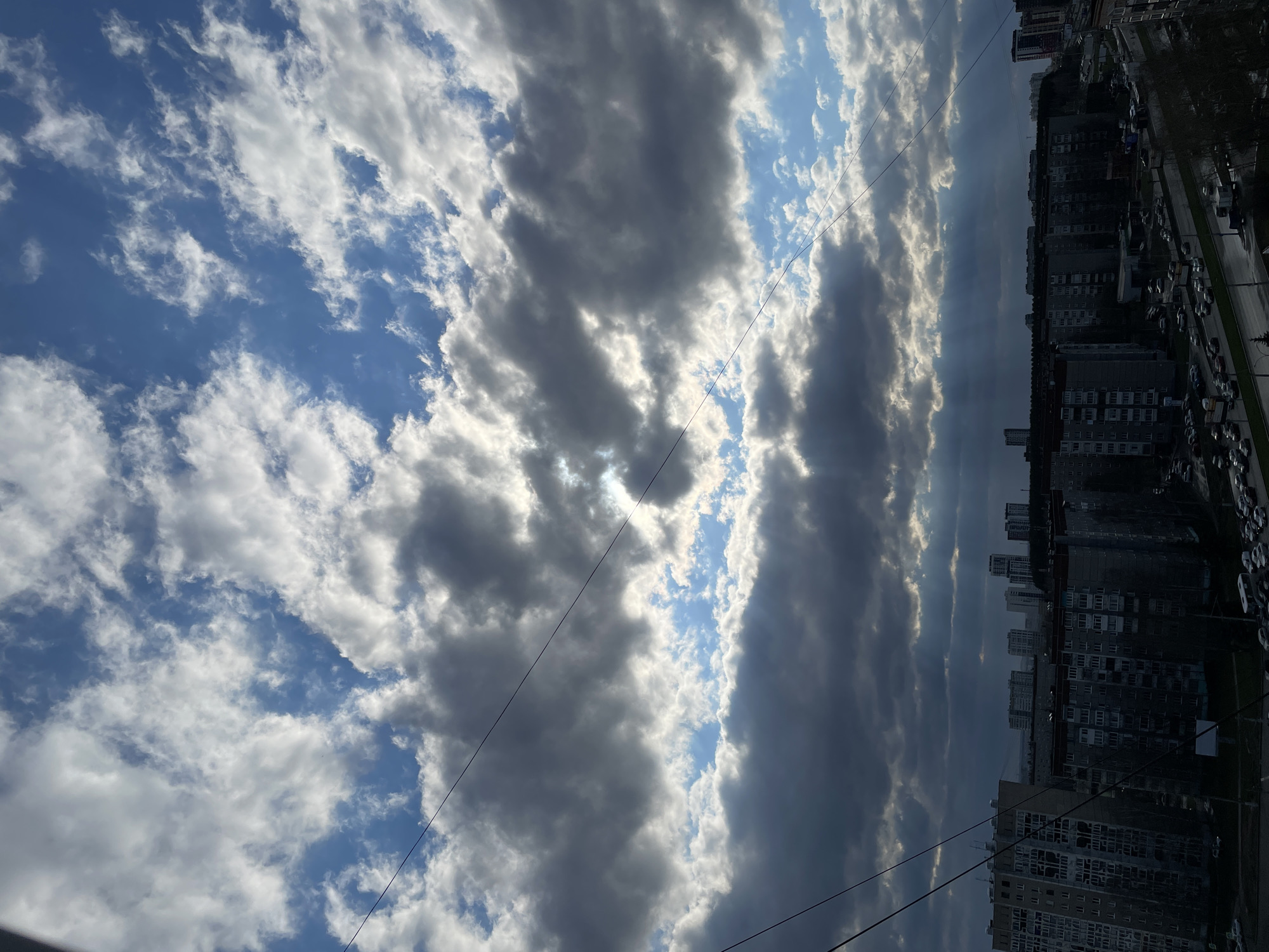 The image displays an urban landscape under a dynamic sky, viewed from a slightly elevated perspective, possibly from a building window. The photo is oriented vertically but depicts a horizontal scene.
The sky occupies the upper two-thirds of the frame and is a striking mix of bright blue patches and various types of clouds. On the left side, there are more scattered, white, fluffy cumulus clouds against a vibrant blue background. Towards the center and right, the clouds become denser and darker, appearing as long, horizontal streaks of grey. Sunbeams (crepuscular rays) are clearly visible breaking through gaps in these darker clouds, illuminating parts of the sky and creating dramatic shafts of light. These rays suggest either early morning or late afternoon light.
Several thin, dark utility or power lines crisscross the sky horizontally in the middle ground.
Below the sky, the lower third of the image reveals a city environment. Numerous multi-story residential or commercial buildings, predominantly in shades of grey, line what appears to be a street or avenue. The buildings have many windows and some show signs of wear. Parked cars are visible along the street and in parking areas adjacent to the buildings. A road with what looks like traffic or more parked cars runs parallel to the buildings. In the distance, more buildings and a faint tree line can be discerned, indicating a sprawling urban area.
The overall impression is one of a typical daytime scene in a city, with strong natural light effects from the sun partially obscured by clouds. The architecture and urban layout are consistent with the provided context of Izhevsk, Russia. No specific activities or visible text are discernible.