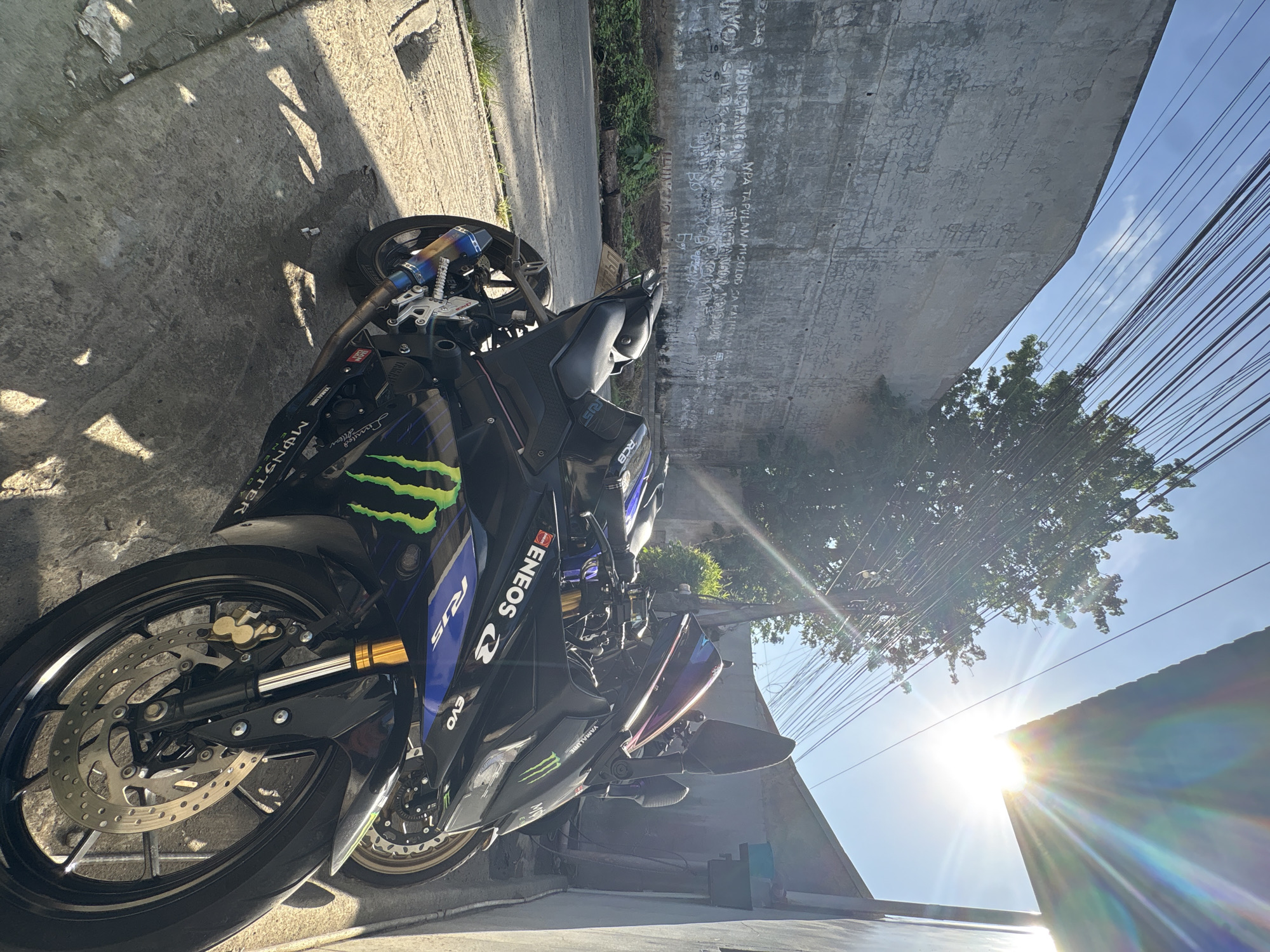 The image depicts a black Yamaha R15 motorcycle parked on a concrete sidewalk in an urban setting, likely in Davao City, Philippines, suggested by the graffiti on the wall. The motorcycle is adorned with racing decals, including "Monster Energy" and "Eneos". Behind the motorcycle, a weathered concrete wall with extensive graffiti, including some text in what appears to be a local dialect, runs along a road. Lush green vegetation grows at the base of the wall and along the roadside. The scene is illuminated by bright sunlight, creating strong shadows and a sunburst effect from the upper right corner of the frame. Overhead, a network of power lines crisscrosses the sky. Another motorcycle is partially visible parked behind the R15. The overall environment appears to be a street in a residential or commercial area of the city.