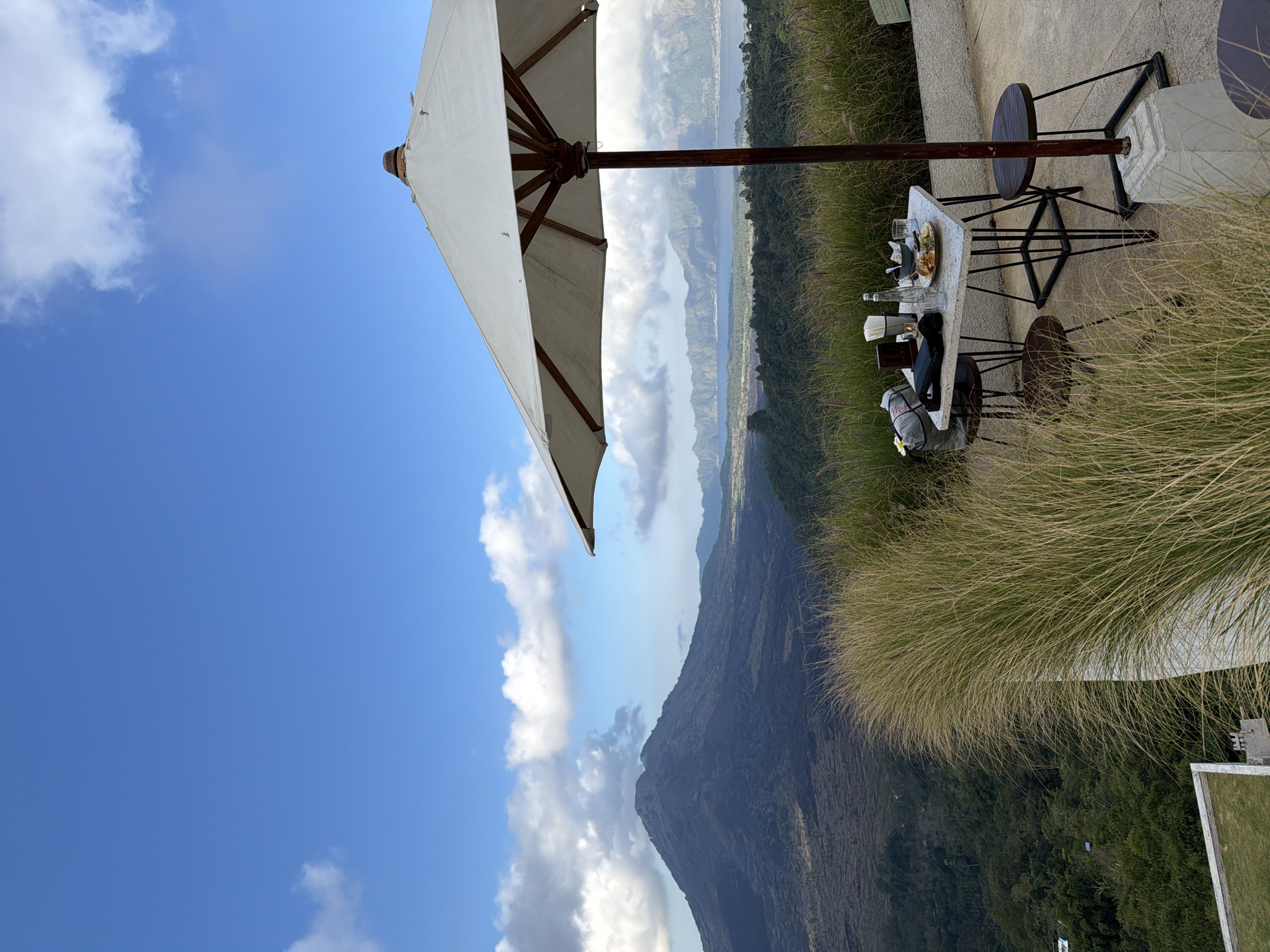 A table for two is set with plates of food, glasses, and napkins under a large white umbrella on a patio overlooking a volcanic landscape. In the background, Mount Batur, a volcano in Kintamani, Indonesia, looms under a blue sky with scattered clouds. The foreground is filled with tall, dry grass. A black bag and a small wooden frame are on the table. Two chairs are placed at the table.