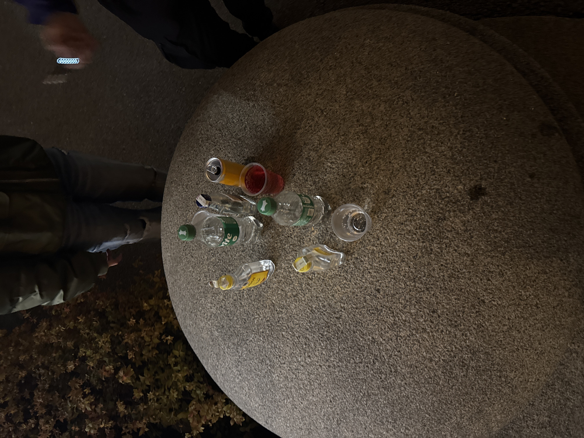 The image captures an outdoor scene at night, centered on a large, round, granular grey-brown stone surface functioning as a table. Scattered across this surface are various empty or partially consumed beverage containers. These include an orange soda can, a clear plastic cup holding red liquid, three small clear plastic bottles (one with a green cap and partial text "rite", likely Sprite), two small clear plastic miniature bottles with yellow labels (one showing "KOY"), and another empty clear plastic cup.
Portions of at least two individuals are visible around the table. In the upper left, a person in a dark jacket holds a device emitting a blue, chain-link pattern of light, possibly a phone or a light-up accessory. To the lower left, another person wears a green or khaki-colored jacket. The ground beneath the individuals appears to be dark asphalt or pavement, and dense shrubbery or bushes are visible in the bottom left corner. The dim lighting confirms it is nighttime, suggesting a casual outdoor gathering or social event. No specific landmarks identify the location as Sofia, Bulgaria.