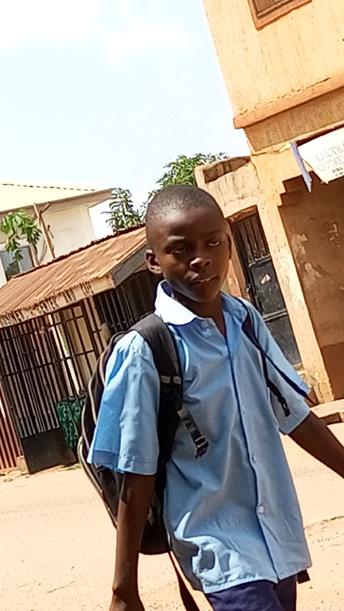 A young boy in a light blue school uniform with a black backpack stands outdoors in Sabon Tasha, Nigeria. He is looking towards the camera. He is surrounded by buildings and trees. The buildings are a reddish-brown color with grey roofs.