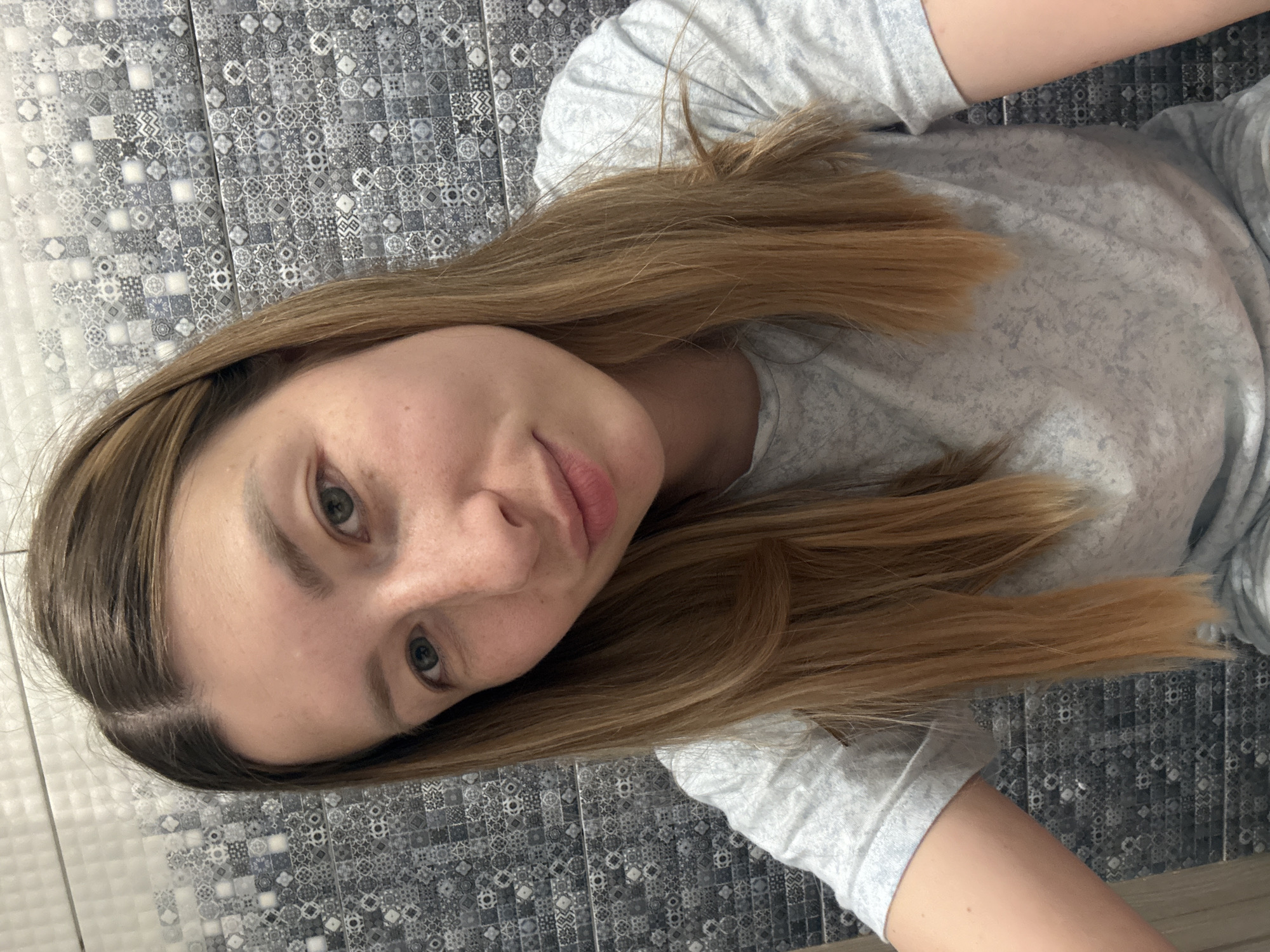 A woman with light brown hair and pale skin is lying on a surface covered in gray and white patterned tiles, possibly a floor or wall. She is wearing a light gray t-shirt. Her gaze is directed towards the camera. The location is unknown and cannot be determined as Moscow, Russia.