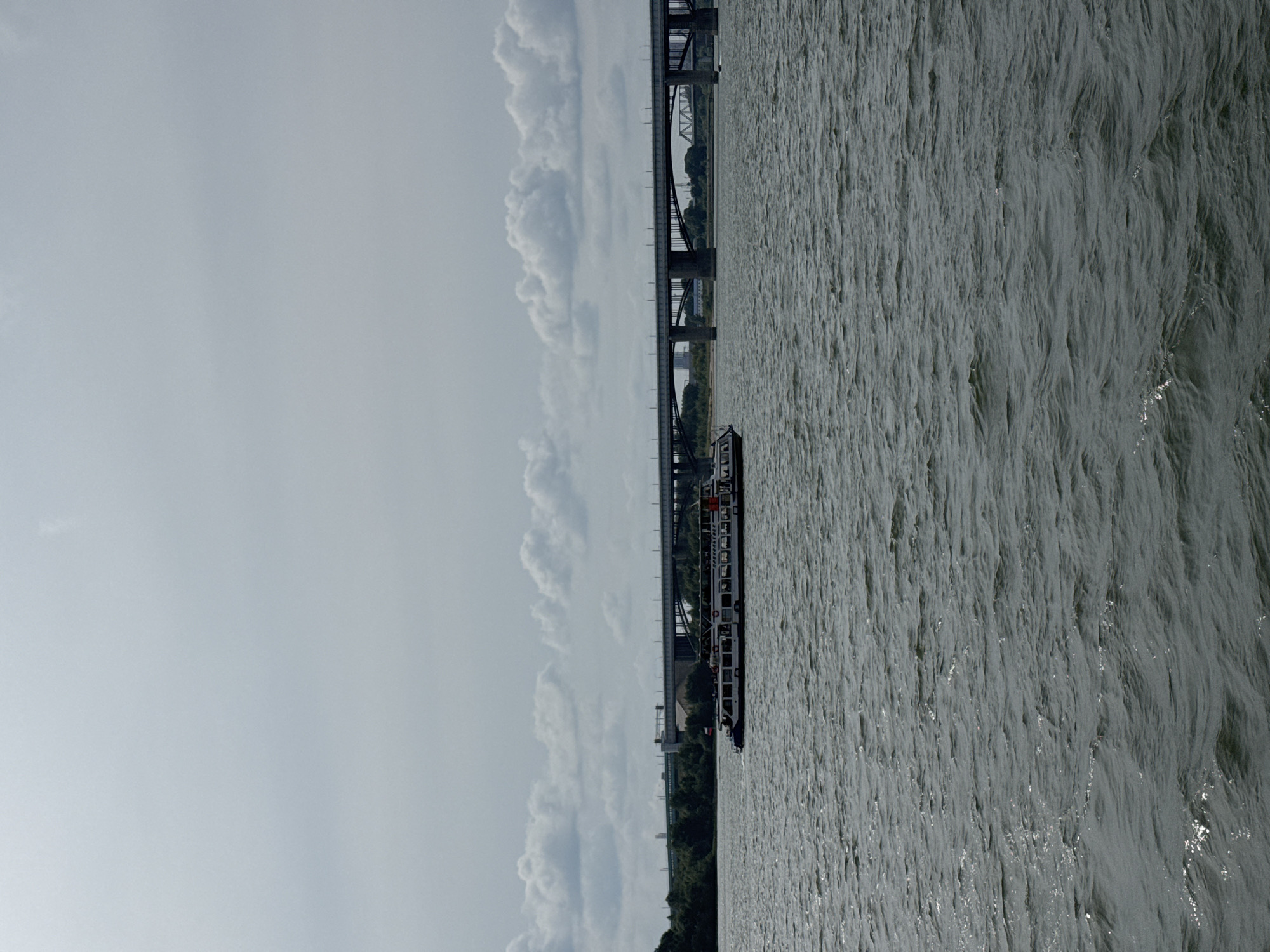 An overcast, daytime, wide shot captures a broad, dark gray river with visible ripples in the foreground. A long, white passenger boat with a dark blue hull travels from left to right across the frame, appearing to be in motion. It has several decks with windows and people can be seen on the upper deck. Behind the boat, a densely wooded shoreline with scattered buildings is visible. Further in the background, a series of large, gray bridges span the river, with an arched bridge prominent in the center. The sky is filled with thick, white clouds. This scene is likely the Ob River in Novosibirsk, Russia, given the presence of large river bridges typical of the city.