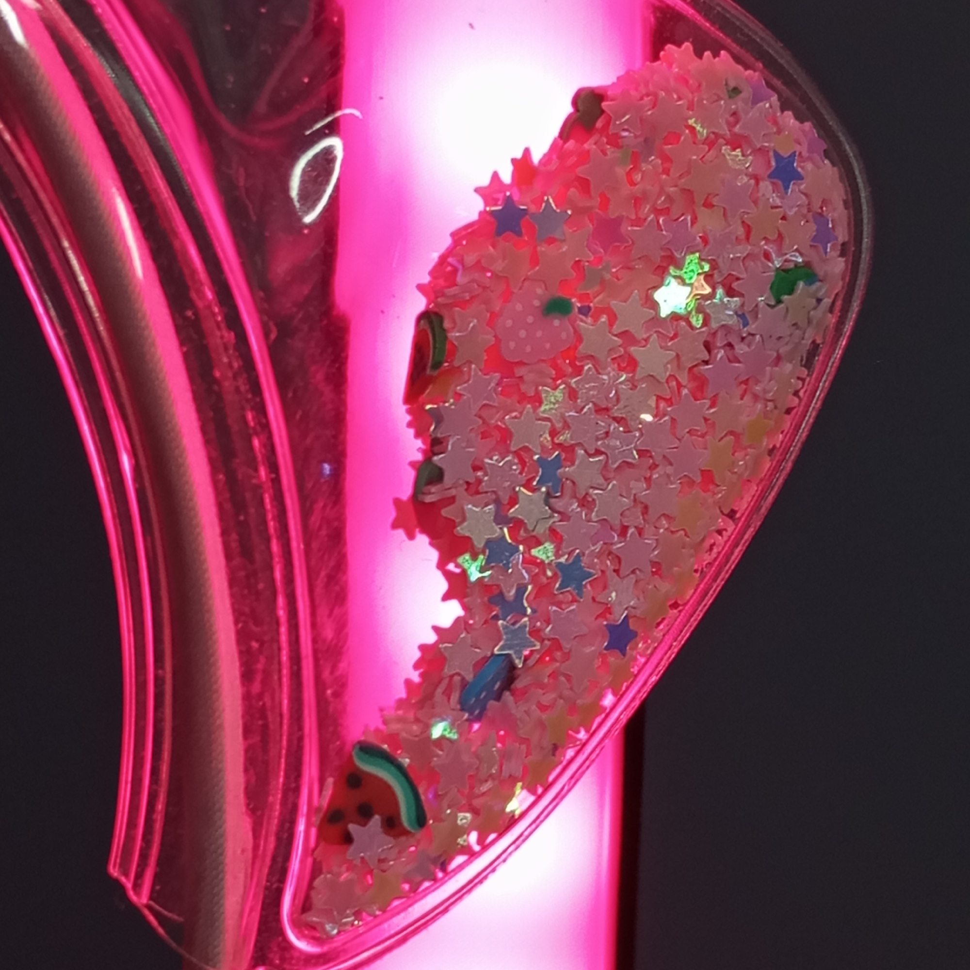 The image is a close-up, high-angle shot focusing on a decorative object with vibrant pink illumination against a dark, indistinct background. The primary object appears to be a clear or translucent container, possibly made of plastic, which is deeply tinted pink. This container has a curved, almost heart-like shape on its right side, holding a collection of small, colorful decorative elements. The left side of the container shows multiple parallel ridges or grooves, indicating a textured or shaped surface.
Inside the clear, pink-tinged vessel, there is a dense cluster of various miniature adornments. The most prominent are numerous star-shaped pieces of glitter or confetti, predominantly in shades of pale pink, cream, or translucent white, with occasional pieces in blue, purple, and iridescent green reflecting the light. Mixed among the stars are distinct fruit-shaped pieces, including a small, pink strawberry with white polka dots and a green stem, and a slice of watermelon with a red interior, black seeds, and a green rind. A small, thin, blue rectangular piece is also visible.
A strong, bright pink light source illuminates the object from behind and within, casting a glow that accentuates the pink hues of the container and some of the contents. The background is uniformly dark, suggesting the photo was taken indoors or in a dimly lit environment. There are no people or specific activities depicted. No text is visible in the image. The setting provides no discernible clues to the specified location of Pervomaysky, Russia.