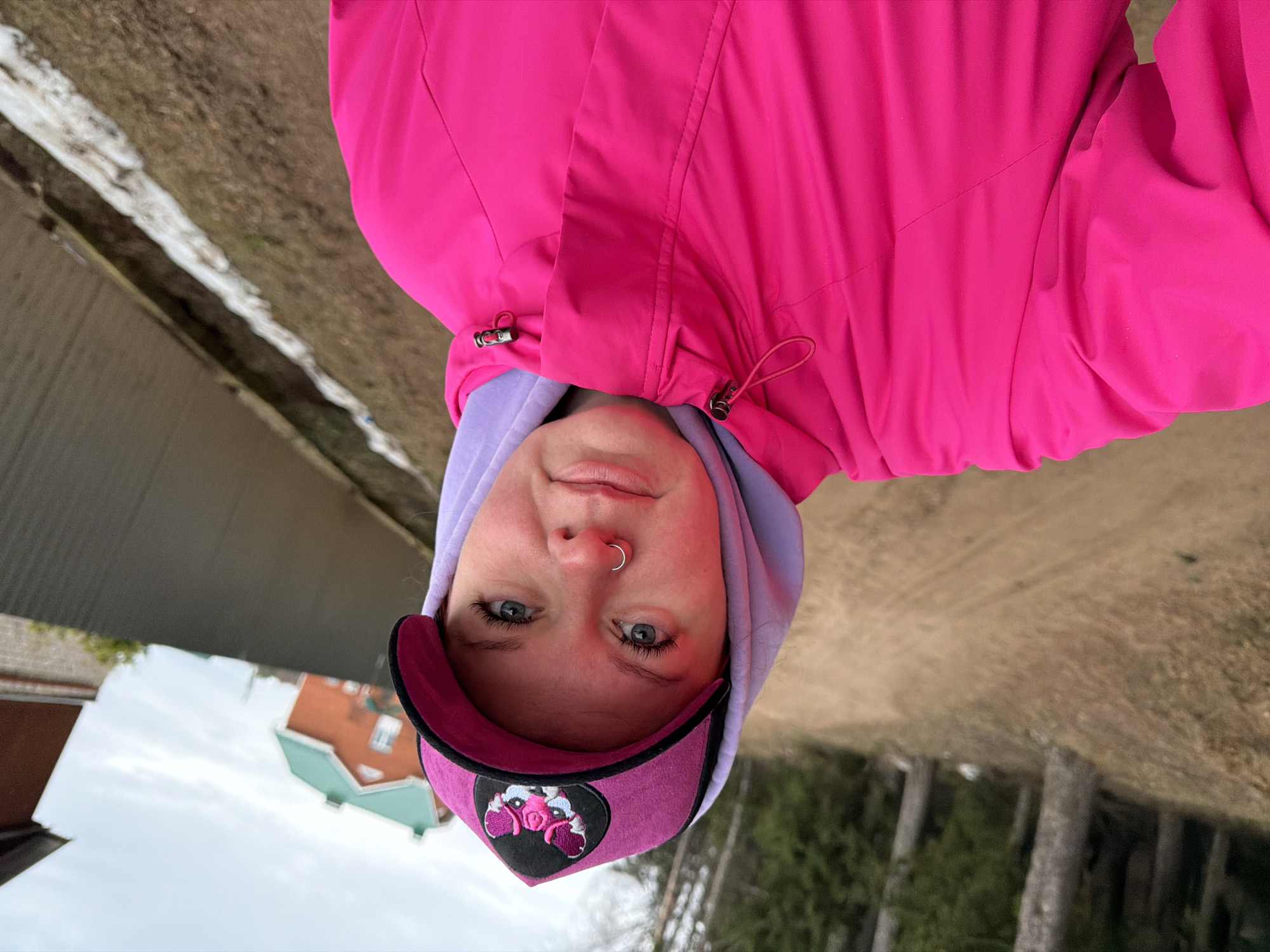 A person wearing a pink jacket, purple hoodie, and pink cap stands outdoors. The background includes trees, grass, and a building with a green roof. The location is Chaikovsky, Russia.