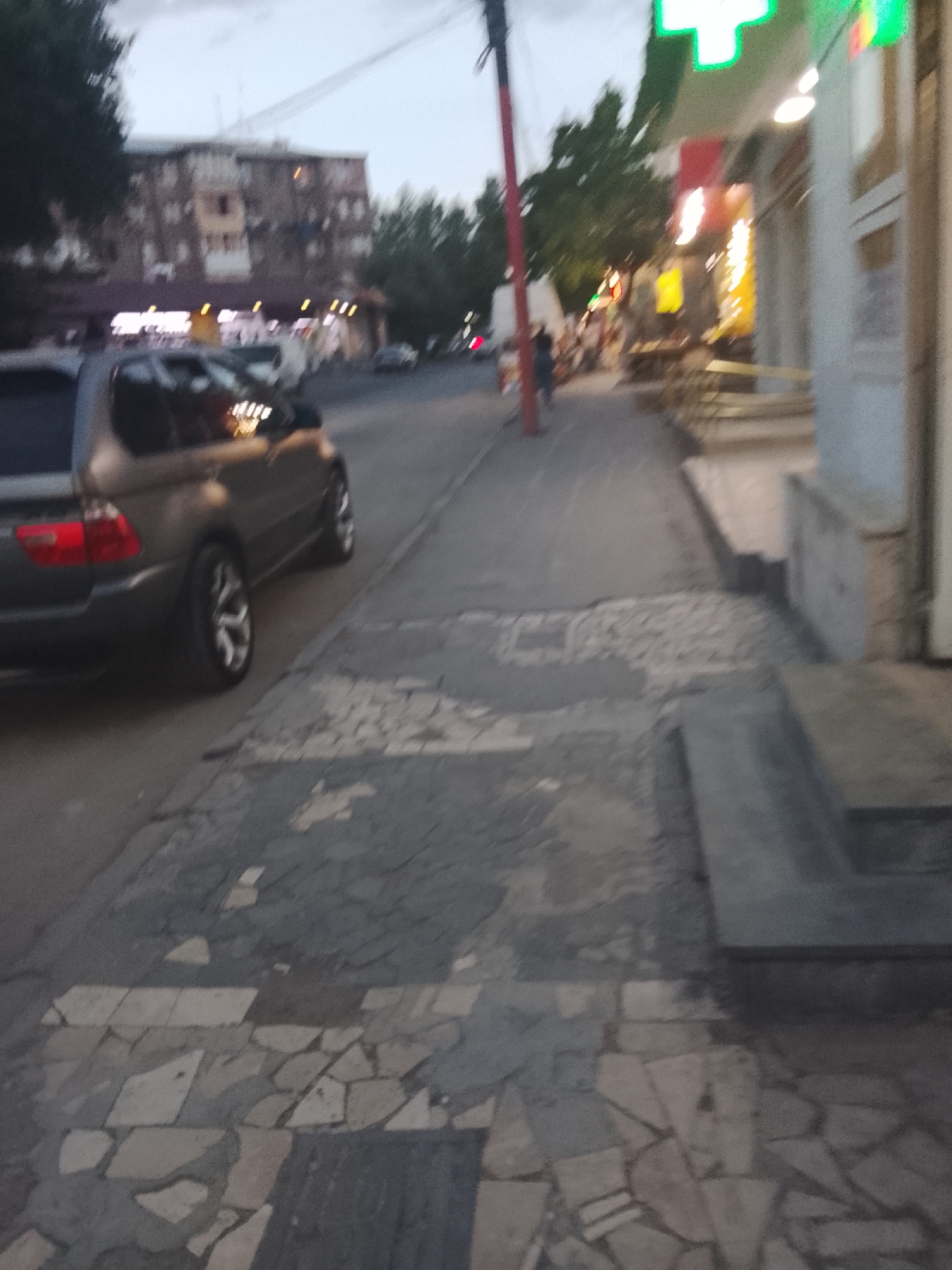 This media file depicts a street scene at dusk. The foreground is dominated by the side of a dark-colored SUV, with its taillights visible. The pavement is made of irregularly shaped stone tiles, some of which are broken or missing. To the right, a building with a brightly lit green cross sign, likely a pharmacy, is visible. Further down the street, there are other buildings, trees, and a few indistinct figures of people walking. The sky is overcast, suggesting a cloudy or possibly rainy day. The overall impression is one of a typical urban environment in the evening. There is no visible text in the media file.