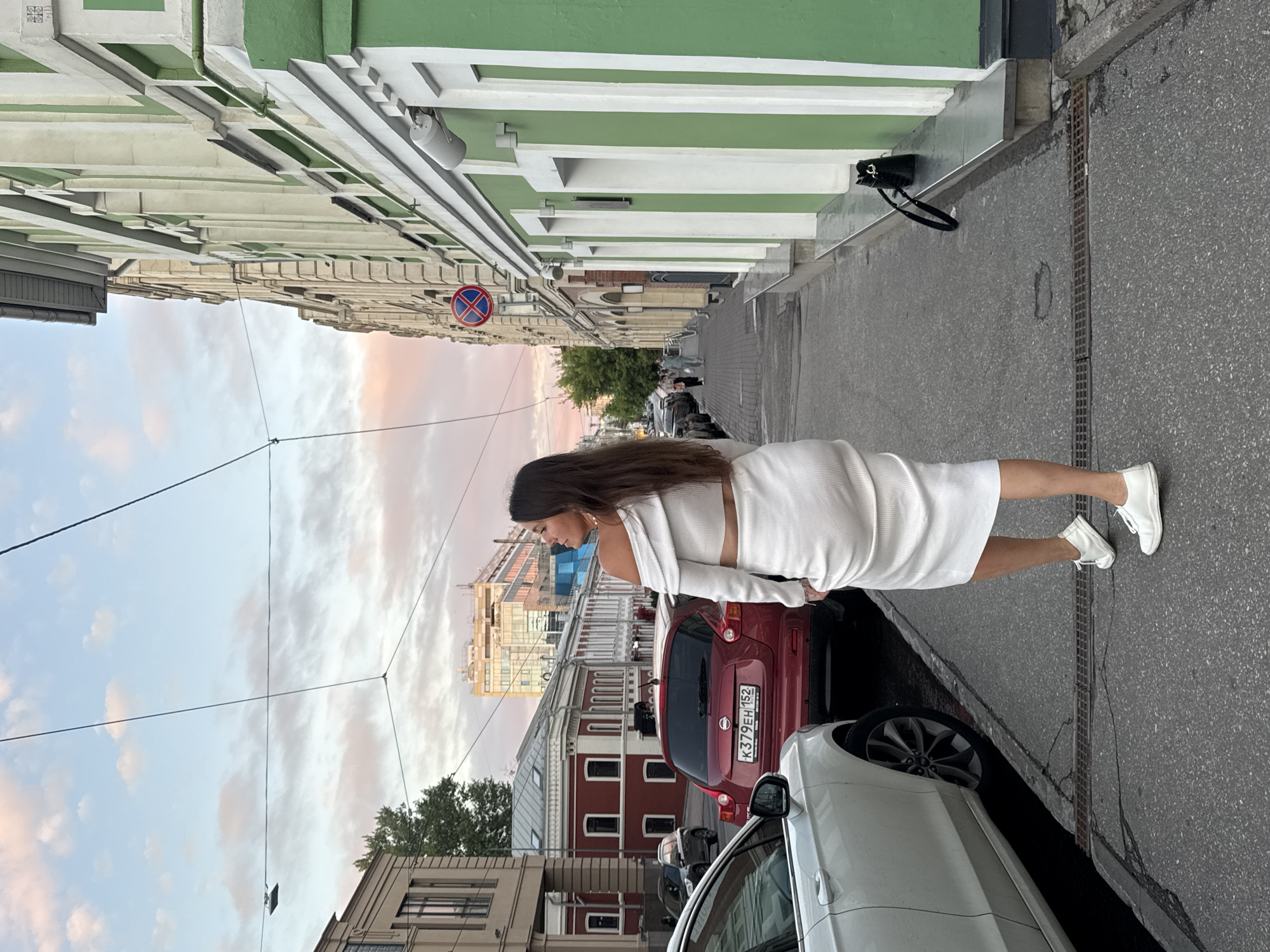 A woman in a white two-piece outfit walks away from the camera on a sidewalk in a city. She has long, dark hair and is wearing white sneakers. She is facing towards a street with cars parked and driving. The buildings on the right are green and tan, while the buildings on the left are red and tan. The sky is a soft pink and blue, indicating sunset. There are power lines running overhead. A red car with a license plate that reads "K379EH 152" is parked on the street. A white car is parked on the left side of the frame. A black purse sits on a ledge in front of a building. The scene appears to be a typical urban environment with residential and commercial buildings. The location is Nizhny Novgorod, Russia.