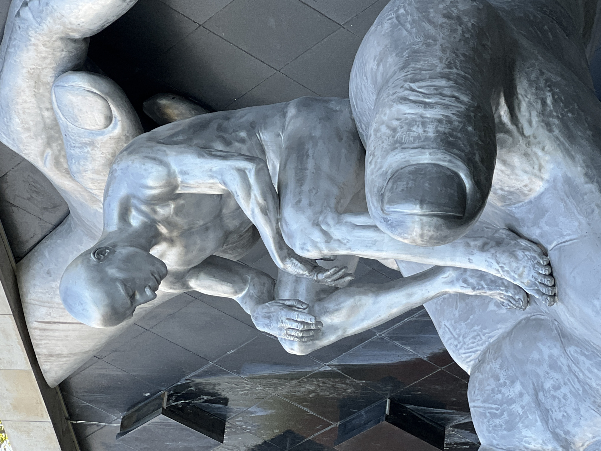 The image shows a sculpture of a bald, nude male figure sitting with his head bowed in contemplation, resting on large, stylized hands. The sculpture is metallic and appears to be silver or pewter in color. It is positioned in front of a dark, tiled wall with a water feature cascading down it. The sculpture is part of the "Hands of Fate" monument, located in front of the House of Architect in Krasnodar, Russia.