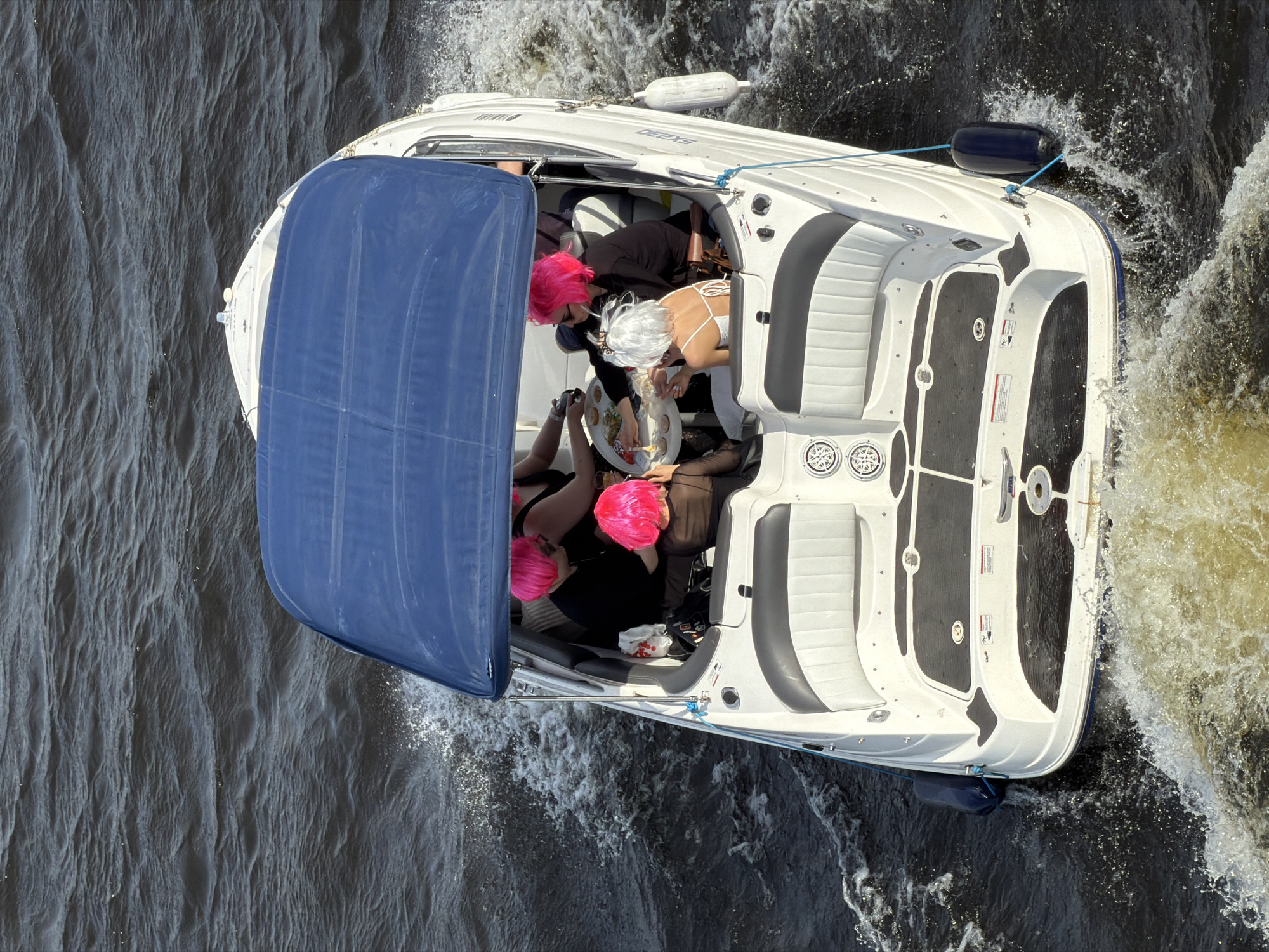 An overhead, high-angle shot depicts a white motorboat navigating dark, choppy water in Saint Petersburg, Russia. The vessel features a white hull, black non-slip surfaces on the deck, and a deployed blue canvas bimini top. Two blue fenders are attached to the boat's port and starboard sides. The boat is in motion, generating a visible wake at the stern and spray at the bow. Inside the cockpit, at least five individuals are present. Four of them are wearing brightly colored wigs: three in vibrant pink and one in white. They are dressed in casual, predominantly dark attire. A small, circular white table in the cockpit holds various items, including glasses containing beverages, small plates, and snacks. Individuals are observed interacting around the table, with one person in a white wig seated facing away from the camera, wearing a white top. The scene is illuminated by daylight.