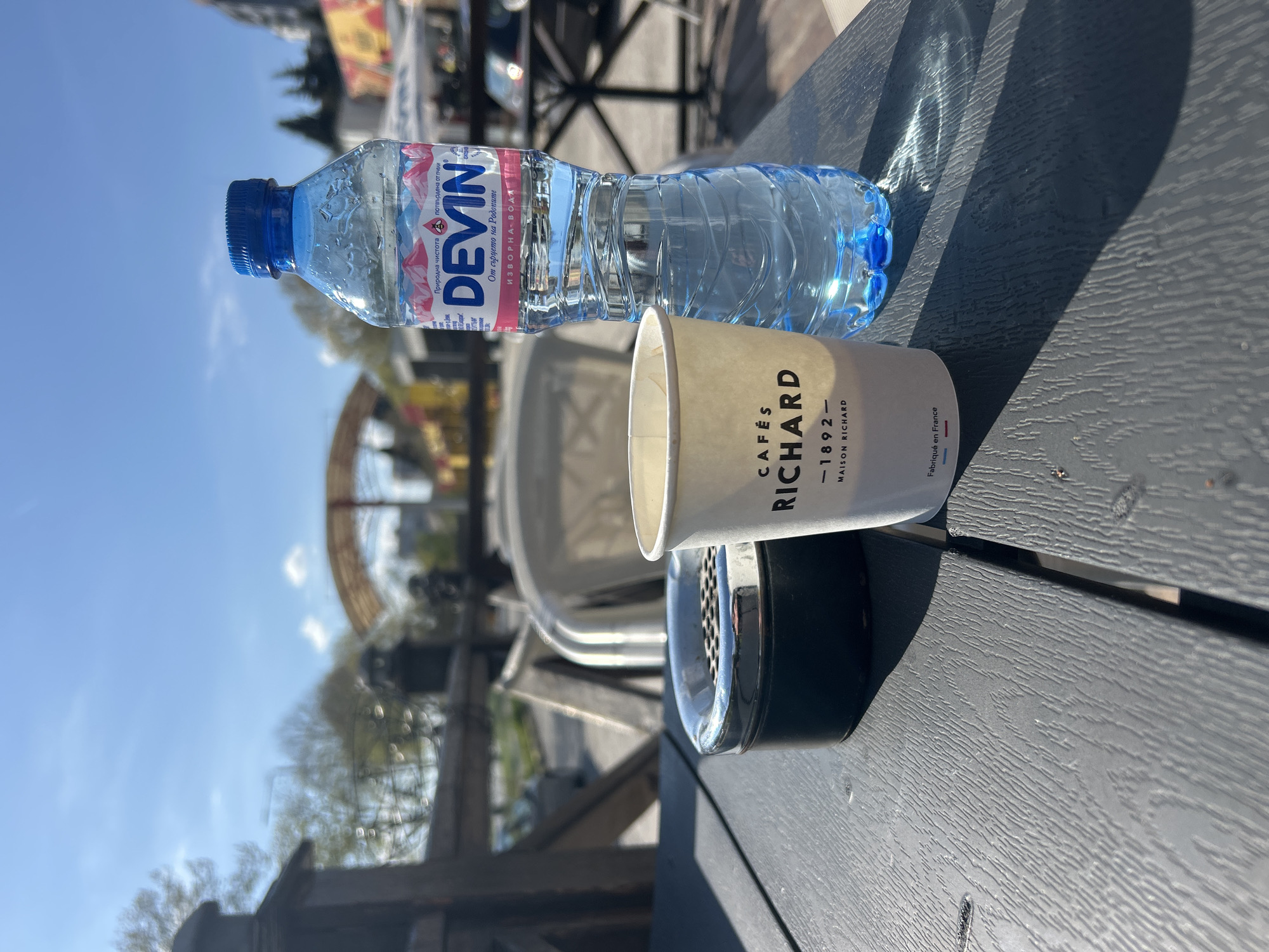 An outdoor scene captured on a sunny day shows a table with refreshments, likely at a café or restaurant terrace. The primary subjects are a plastic bottle of water and a paper cup, resting on a dark grey table with a textured surface.
The water bottle, branded "DEVIN," is filled with clear water and features a blue cap and label. Visible text on the label includes "DEVIN" prominently, along with Bulgarian phrases such as "Природна чистота" (Natural purity), "потвърдена от пчели," "От сърцето на Родопите" (From the heart of the Rhodopes), and "ИЗВОРНА ВОДА" (Spring water).
Next to the bottle is a white paper cup, bearing the brand "CAFÉS RICHARD" with the text "-1892-" and "MAISON RICHARD." Below this, "Fabriqué en France" (Made in France) is visible. A black, round object, possibly an ashtray, is situated beneath the cup.
The scene is brightly lit by the sun, casting long, distinct shadows from the bottle and cup onto the table, suggesting it is either mid-morning or late afternoon. The background is softly blurred but reveals a clear blue sky, some trees, and indistinct architectural elements or structures, indicative of an urban or semi-urban outdoor setting. No people are visible. The presence of the Devin water bottle suggests a location in Bulgaria, despite the coffee cup being from a French brand. The overall impression is one of a relaxed break in pleasant weather.
