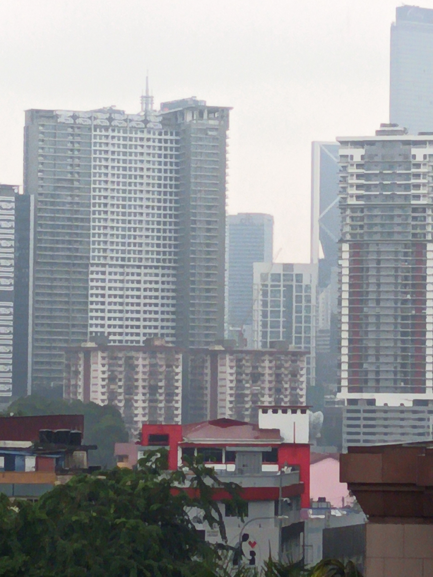 The image depicts a cityscape, likely Kuala Lumpur, Malaysia, characterized by a cluster of high-rise buildings under a hazy, overcast sky. The primary structures are large, rectangular residential or office buildings with numerous windows. Some buildings appear to be under construction or recently completed, as suggested by exposed frameworks and unfinished facades.
In the foreground, there are lower-rise buildings with red roofs and some exposed concrete structures. Lush green foliage is visible in the bottom part of the image, suggesting the presence of trees and vegetation within or around the urban environment. The overall impression is one of a densely populated urban area experiencing overcast weather conditions. There are no discernible people or specific actions taking place within the frame. The scene is static and focuses on the architectural landscape.