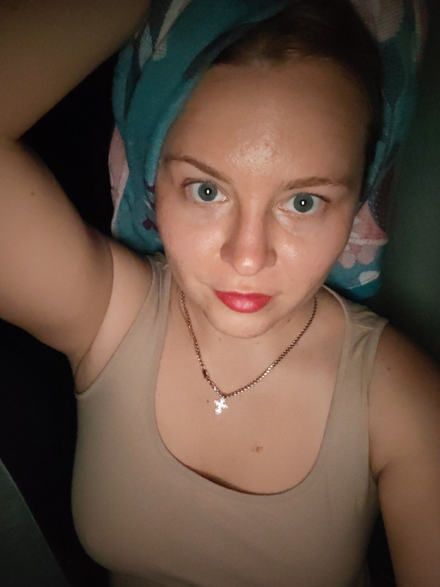 A close-up selfie shows a young woman with fair skin, blue eyes, and red lipstick. She is wearing a beige tank top and a necklace with a cross pendant. A teal and pink patterned towel is wrapped around her head. The background is dark. The image appears to have been taken indoors in Izhevsk, Russia.