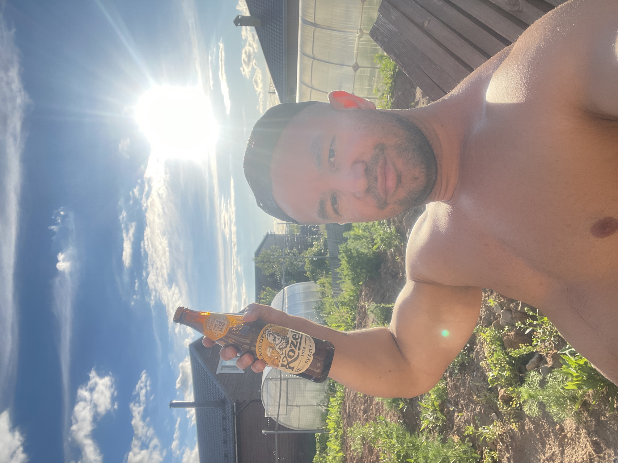 A shirtless man is holding a bottle of Kozel beer in an outdoor setting. The background includes a building with a grey roof, a greenhouse-like structure, and vegetation. The sky is visible with clouds and a bright sun. The bottle is held in the man's outstretched arm, with the label clearly visible.