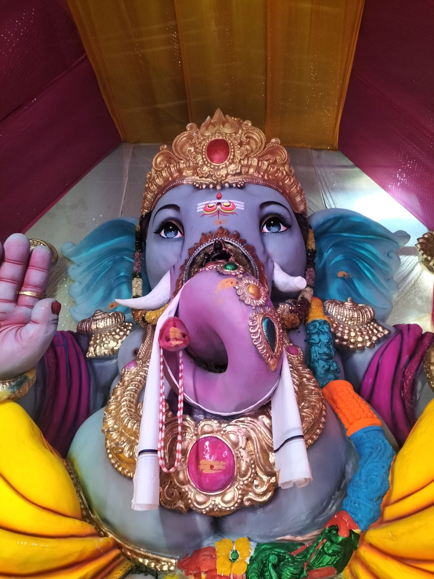 A low-angle, eye-level shot shows a close-up of a large, grey statue of the Hindu deity Ganesha. The statue is adorned with elaborate gold jewelry, including a crown, necklaces, and armbands. Ganesha's trunk is painted purple and is shown curled to the side, with a decorative pink jewel in the center. Ganesha's eyes are large and blue, with black eyeliner and prominent eyelashes. A white cloth is draped around Ganesha's neck. The statue is positioned under a fabric canopy, with swathes of yellow and magenta fabric draped above, creating a festive atmosphere. The background is blurred, suggesting the statue is part of a larger event or celebration. The image was taken in Ishratabad, India.