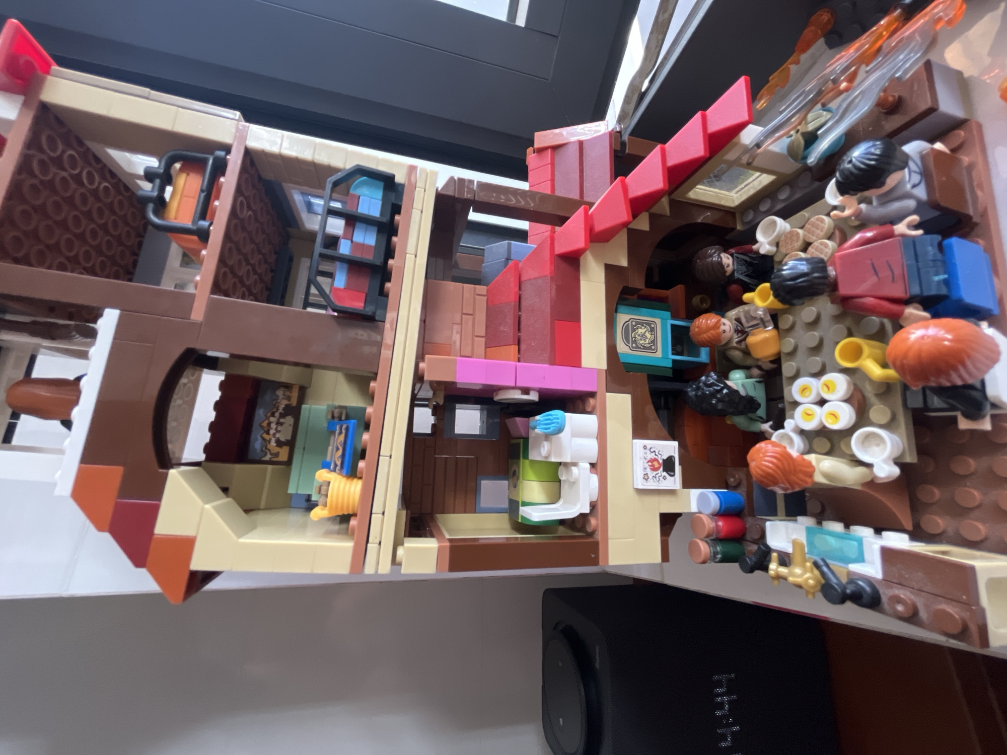 A LEGO model of a building interior is visible in the image. The model has multiple rooms and levels. Miniature LEGO figures are positioned within the rooms, appearing to engage in various activities. The model includes furniture and details suggestive of a domestic setting, such as tables, chairs, and beds. The model is indoors and partially obscures a black smart speaker on a white shelf. The location, Bac Nha Trang Ward, Vietnam is not discernible.