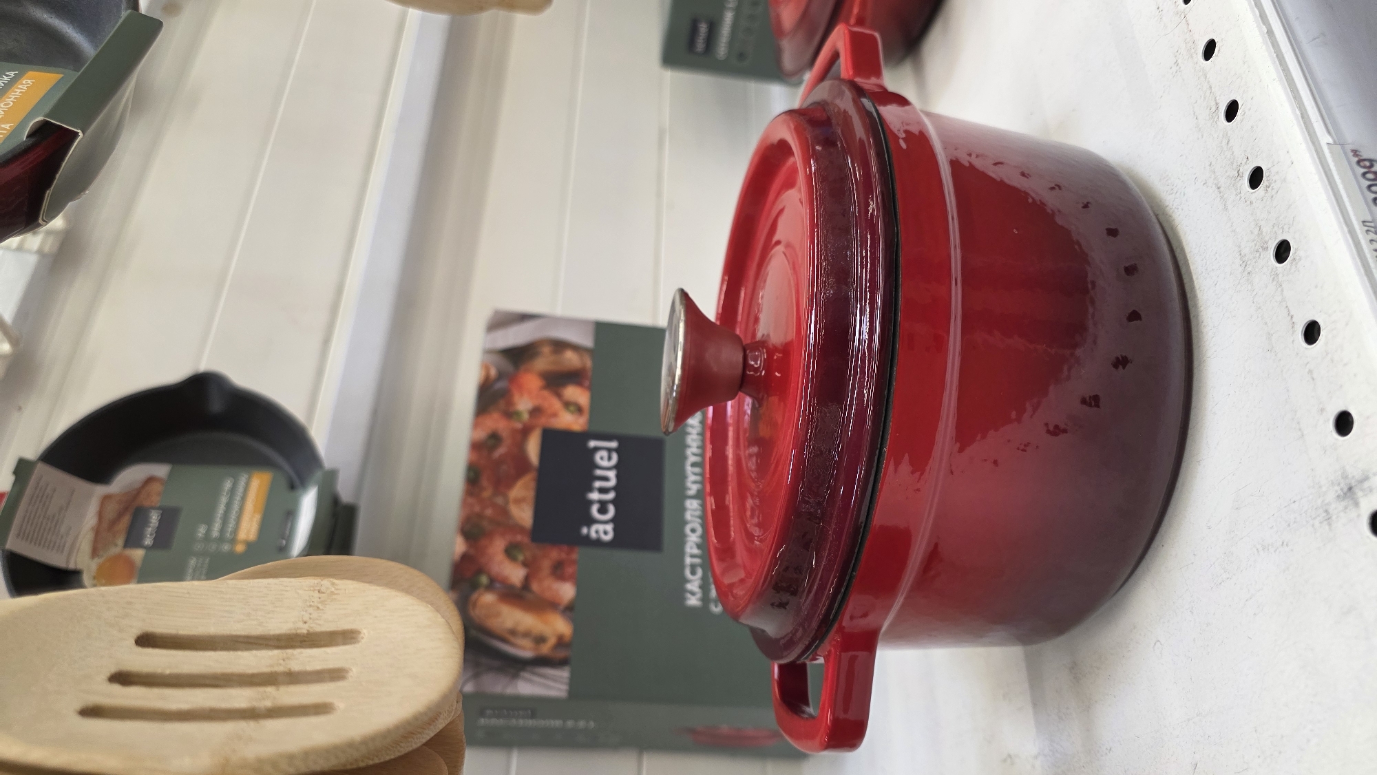 This image, rotated 90 degrees counter-clockwise, depicts a display of kitchenware, likely in a retail store setting. The main subject is a vibrant, glossy red enameled cast iron pot, or dutch oven, prominently positioned in the center. It has a matching red lid with a knob on top and two handles on its sides.
To the left of the red pot, partially visible, are several light-colored wooden kitchen utensils, including a slotted spoon, stacked together. Further to the left and slightly behind, a black frying pan, also in its packaging, is visible.
In the background, behind the cookware, are several product boxes. One box, primarily dark green with an image of cooked food (possibly seafood or meat) and the brand name "actuel," is clearly visible. Text on this box in Cyrillic reads "КАСТРЮЛЯ ЧУГУННАЯ," which translates to "Cast Iron Pot." Another partial label on the far top left also shows "ЧУГУННАЯ" (Cast Iron).
The background consists of light-colored, possibly white, shelves or walls. On the far right, a white shelf edge with a series of black peg holes is visible, typical of retail shelving. A price tag or label is partially seen on this shelf, with "600" and "2.2" legible. The overall impression is a clean, organized display of new kitchen products.