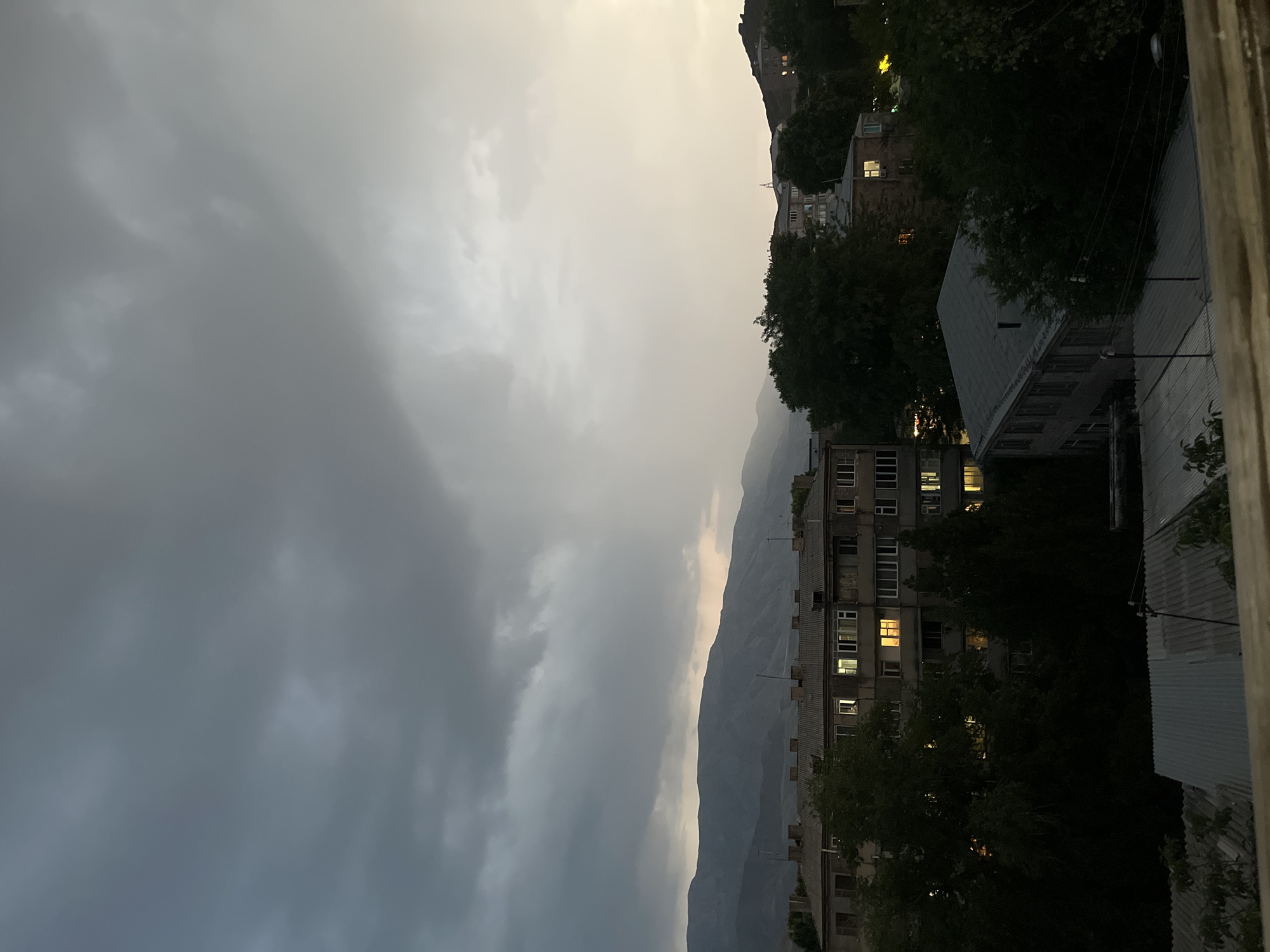 The image depicts a cloudy sky over the city of Gladzor, Armenia. The foreground features the roofs of several buildings, some with corrugated metal coverings. Behind these structures, an apartment building with numerous windows, some illuminated, is visible. Trees surround the buildings, some appearing dark and dense. In the background, a range of mountains stretches across the horizon. The overall lighting suggests it is either dawn or dusk, with the sky being overcast and a soft, diffused light present. There are no people or animals visible in the scene.