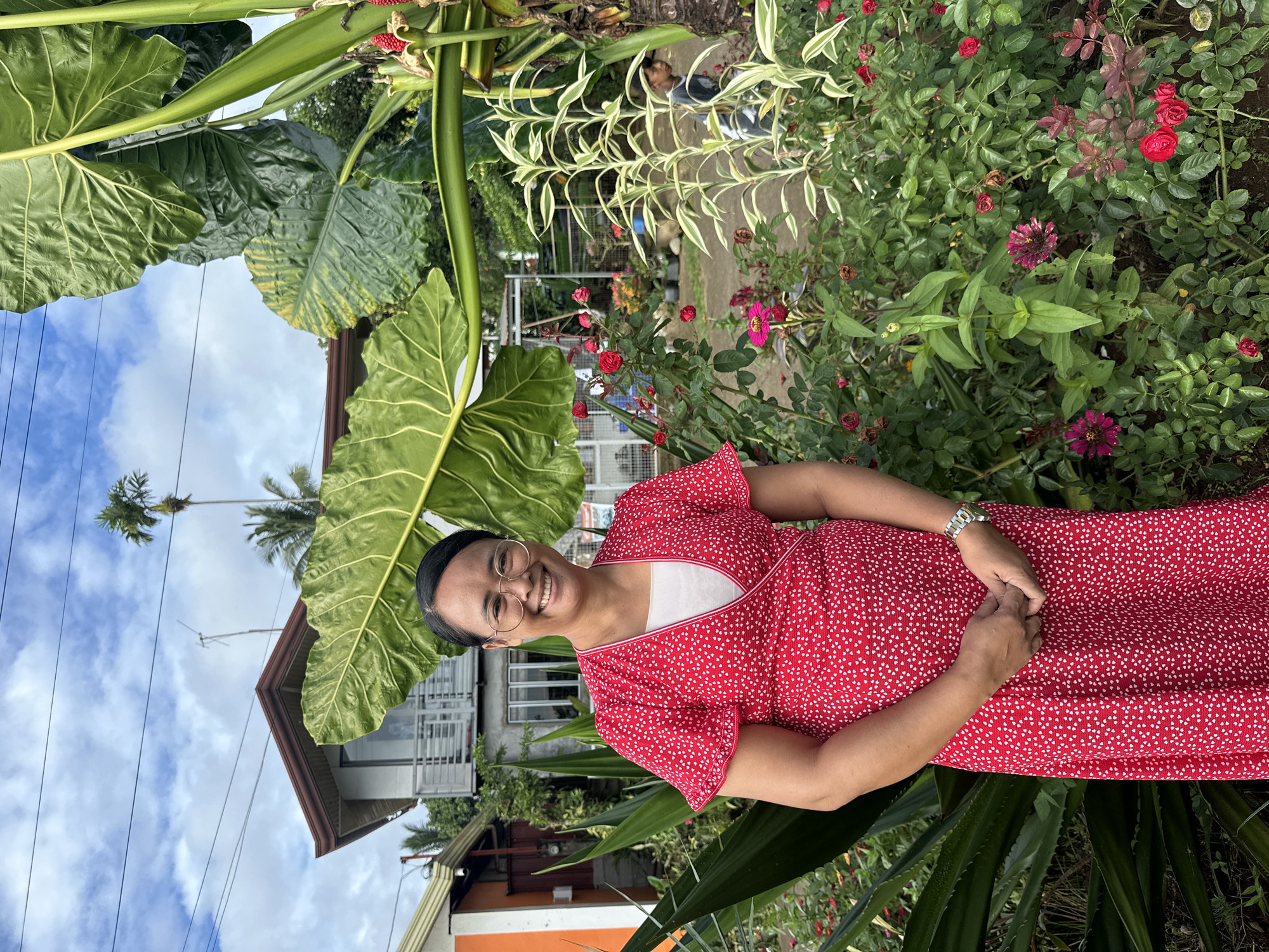 A woman in a red dress stands in a garden. She is smiling and has her hands clasped in front of her. Behind her is a lush garden with a variety of plants, including leafy greens and red flowers. In the background, there is a house with a brown roof and white walls. The sky is visible above the house, with blue patches and white clouds. Based on architectural style, the setting likely is in Davao City, Philippines.