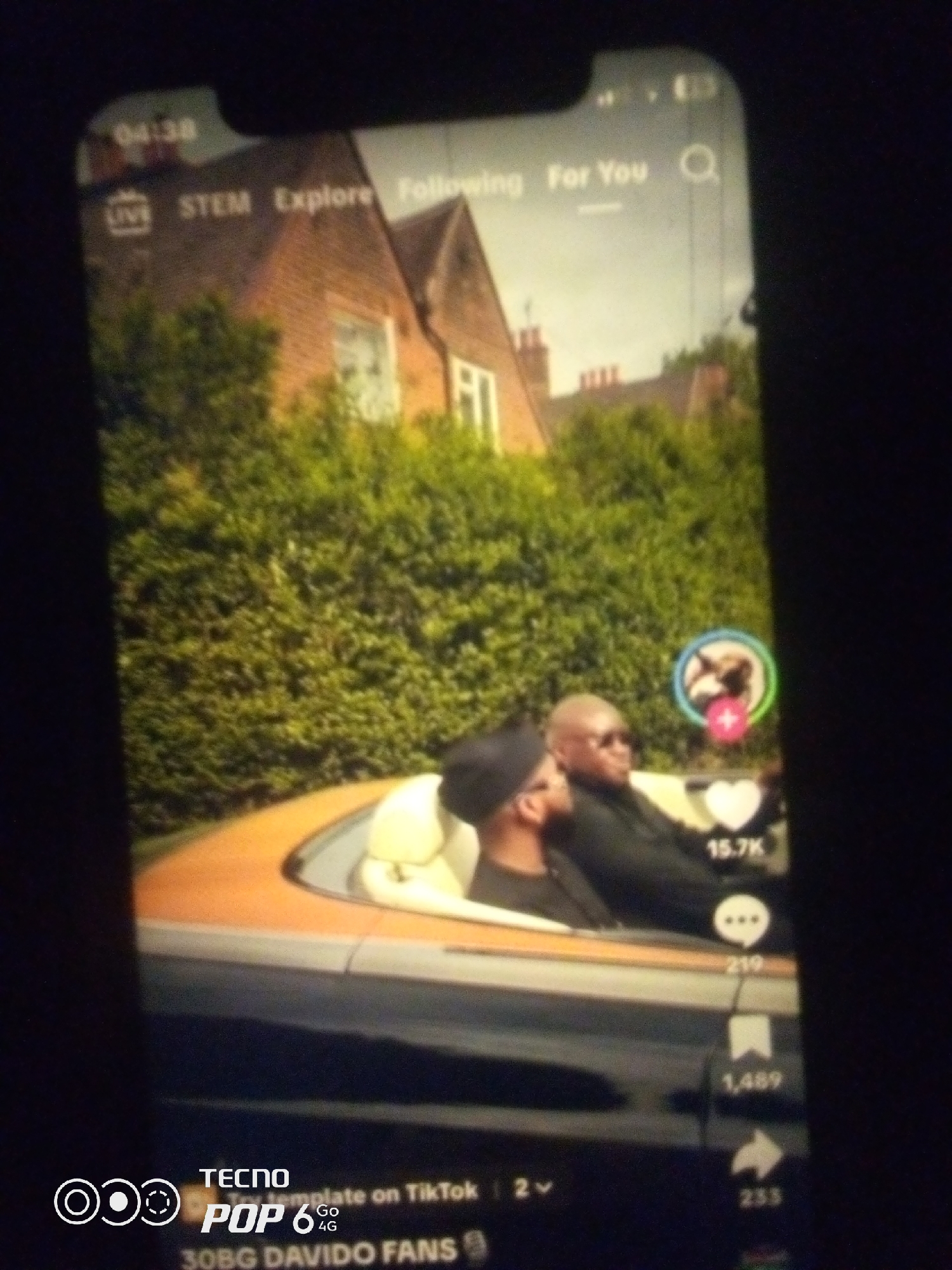 The image shows a TikTok video playing on a smartphone screen. The video depicts two men in a convertible car. One man, in the passenger seat, is wearing sunglasses and a black t-shirt. The driver, partially obscured, is wearing a black beanie. The car is driving past a lush green hedge and a brick house with a red tiled roof. The video interface shows "04:38" at the top, along with "LIVE STEM Explore Following For You". Social media engagement metrics such as "15.7K" likes, "219" comments, "1,489" shares, and "255" views are visible on the right side of the screen. The bottom of the screen displays "TECNO POP 6 4G" and "30BG DAVIDO FANS". While the image shows a scene that could be in Nigeria, there is no specific visual information to confirm the location as Howayi, Nigeria.