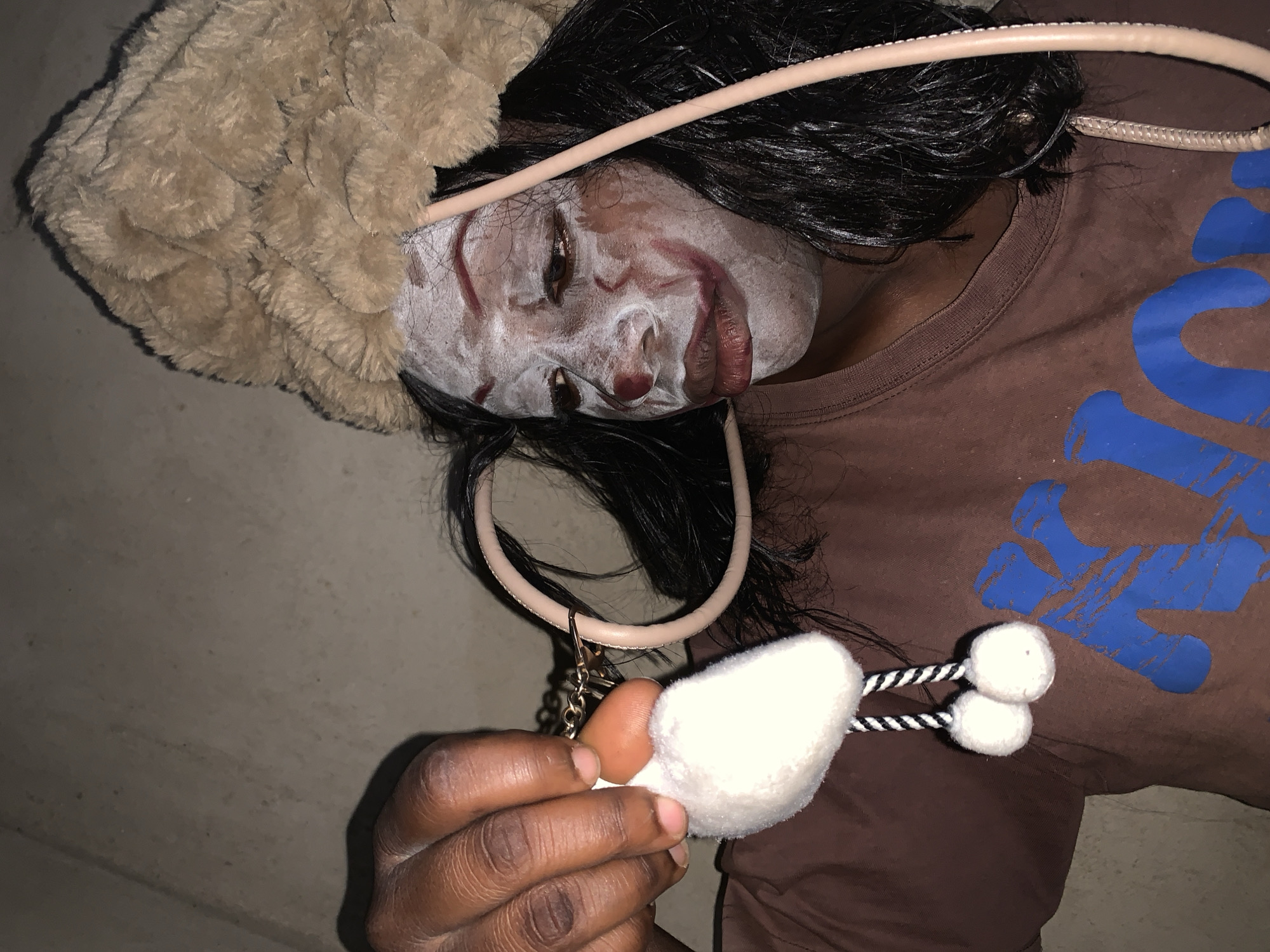 A medium shot of a dark-skinned person lying on a light gray surface. The person is facing away from the camera, with a brown t-shirt visible. There is a large, blue design on the shirt. The person's face is covered in white makeup with red streaks. The person has black hair and is wearing a beige fuzzy hat. The person is also wearing a beige choker and holding a white and black toy. The location of Agban, Nigeria cannot be determined from this image.