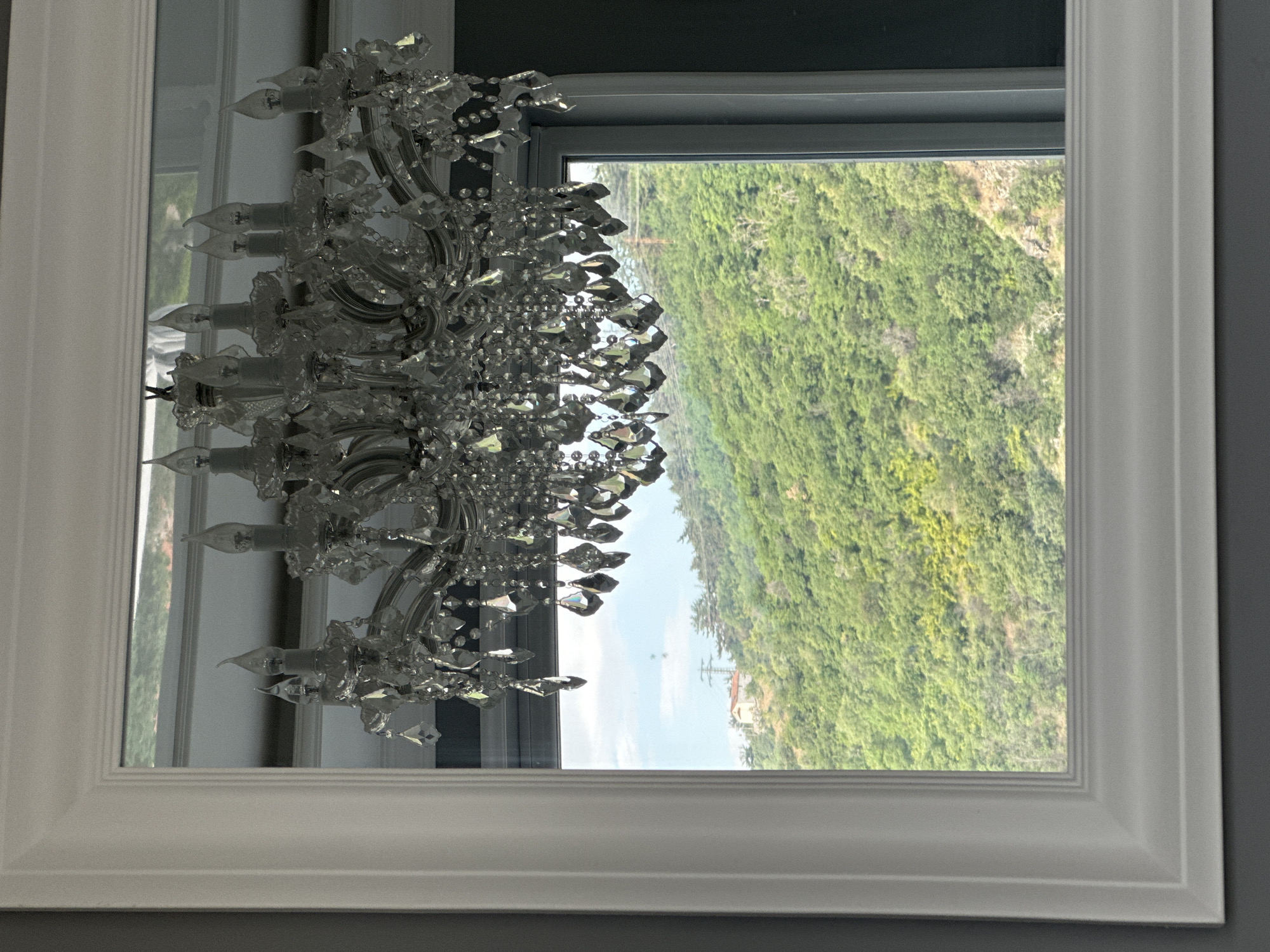 The image displays an interior perspective looking out through a white-framed window onto a verdant, mountainous landscape, specified as being in the city of Asenovgrad, Bulgaria. In the immediate foreground, a multi-tiered crystal chandelier hangs centrally, its numerous faceted drops and candle-shaped bulbs catching the light and partially obscuring the upper portion of the view. The chandelier's frame is a metallic silver. The window itself is framed by a thick, white, decorative molding, which is set against a dark gray or charcoal-colored interior wall on the left and right sides.
Beyond the window, a steeply sloped hillside is densely covered with lush green trees and vegetation, suggesting a thriving natural environment. The foliage appears mature and varied in texture. Towards the upper left horizon of the hill, a small, light-colored building, possibly a house with a reddish roof, is visible. Adjacent to this structure, a dark utility pole with visible power lines extends upwards, indicating human infrastructure within the natural setting. Above the hillside, a clear blue sky with sparse white clouds is present. No people or discernible actions are depicted in the scene.