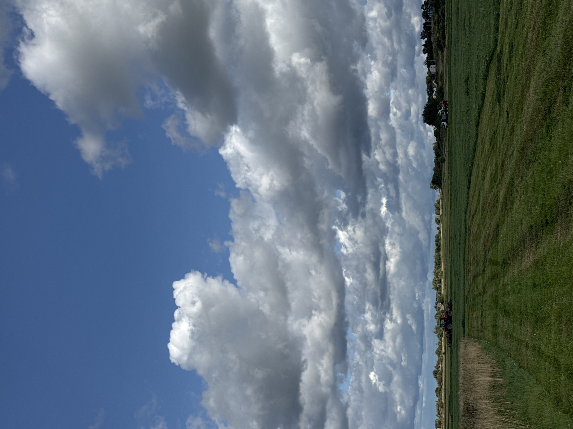 The image captures a wide view of a rural landscape under a dramatic sky. The sky is a vibrant blue, interrupted by large, billowing white and grey clouds that cast shadows on the scene below. In the foreground, a vast expanse of green grassland stretches out, with neat rows of cut grass visible in some areas, indicating recent agricultural activity. To the left, a patch of dry, tall grass borders the mown field.
Further in the distance, a red tractor is actively working in the field, cutting grass. Beyond the fields, a line of trees and scattered buildings marks the horizon, suggesting a small settlement or farmsteads. Another vehicle, possibly a truck, is also visible near the tractor. The overall impression is one of agricultural work taking place in a picturesque, open rural setting. The location is identified as Kempelevo, Russia.