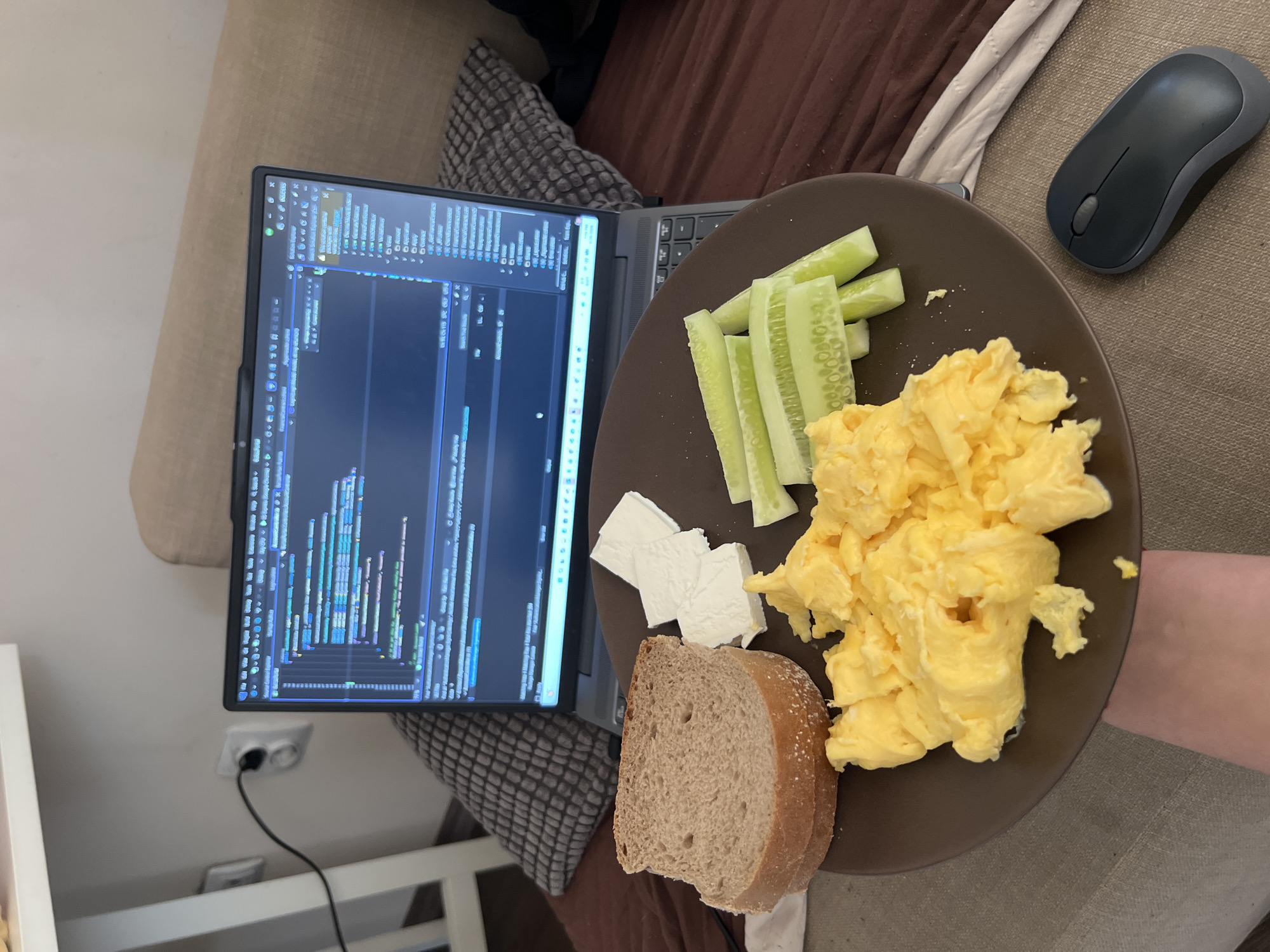 A person is working on a laptop with code displayed on the screen. In front of the laptop, a dark brown plate holds a breakfast consisting of scrambled eggs, slices of white cheese, sliced cucumbers, and a slice of bread. A dark gray computer mouse is placed on the sofa next to the plate. The setting appears to be a living room, likely in Sofia, Bulgaria, given the electrical outlet visible on the wall, which is typical for European standards. The laptop is plugged into the outlet, suggesting the person is settled in for a period of work. The overall scene conveys a sense of focus and productivity, with a balanced meal at hand. There is no visible text in the image.