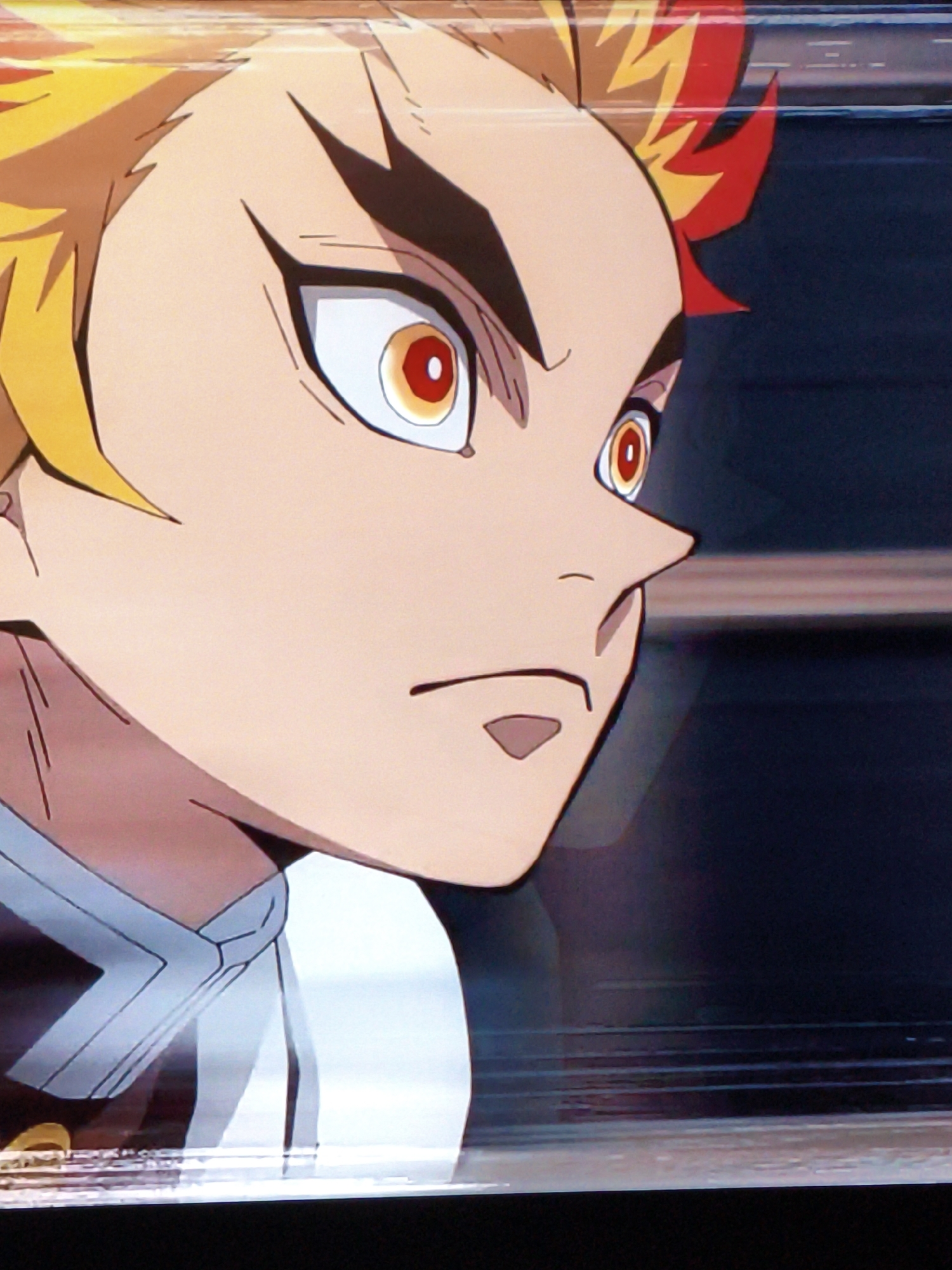 The image is a close-up, medium shot of an anime character's face, viewed from the side. The character has bright yellow and red spiky hair, with prominent black eyebrows arching over large, red-and-yellow eyes. The character's skin is a light peach color. The character is wearing a white uniform with light grey trim around the collar and chest area. The background is dark and appears to be blurred, with horizontal lines suggesting movement or a speed effect. There is no discernible connection to the city of Vladivostok, Russia, or any specific urban environment. The image is a still from an animated work.