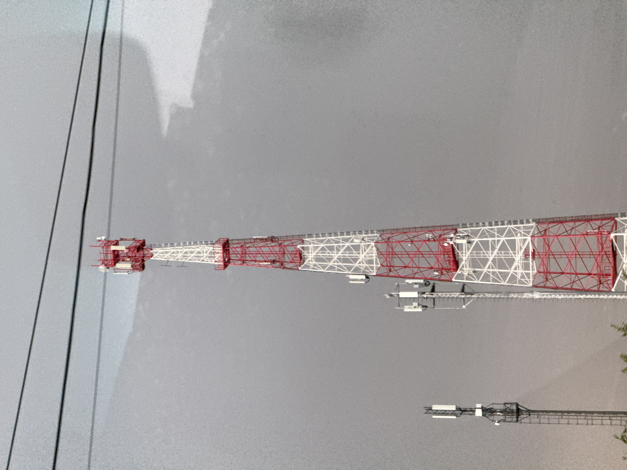 The image shows a red and white lattice radio tower in Malaya Purga, Russia. The tower is seen against a gray, overcast sky. Cables are visible near the top left corner. A smaller, darker tower is visible towards the lower right, partially obscured by foliage.