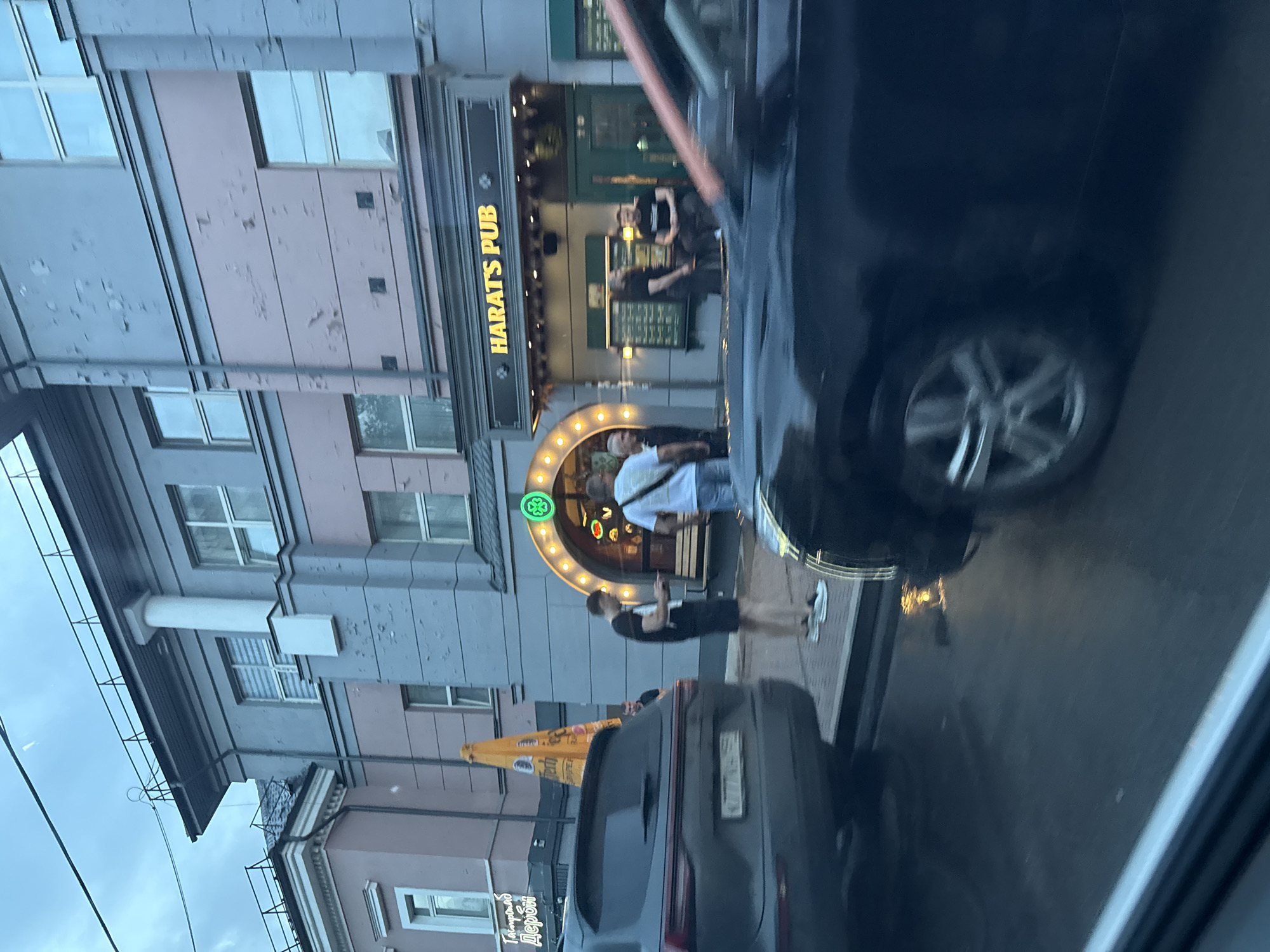 The image captures a street scene in Perm, Russia, on an overcast day, possibly after rain, as indicated by the wet pavement and reflections. In the foreground, a dark-colored car is partially visible, with its wheel and side in focus. The middle ground features a row of buildings. The most prominent is "HARAT'S PUB," a establishment with a distinctive arched entrance adorned with lights and a green shamrock logo. Several individuals are visible near the pub entrance; one man in a black t-shirt and shorts is looking at his phone, while others stand and converse. A dark SUV is parked to the left of the pub, with a license plate partially obscured. An orange umbrella-like structure is also present. The building facades show signs of age, with some peeling paint. The overall impression is a casual urban setting, likely an evening or late afternoon.