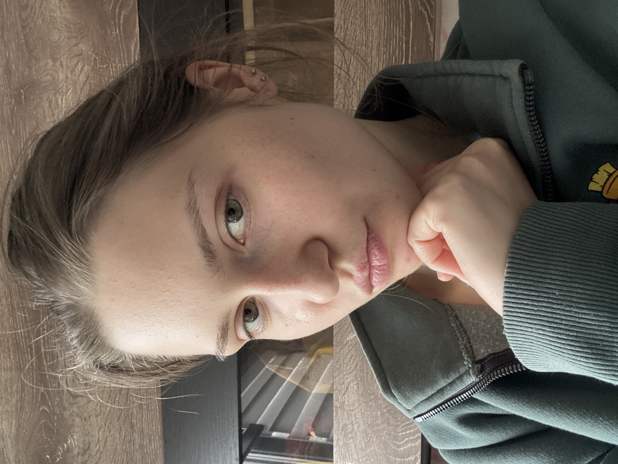 A young woman with light complexion is shown in a medium close-up self portrait. Her eyes are a hazel or green color. She appears to have pierced ears with small stud earrings. She's wearing a dark green zippered sweatshirt. Her left hand is placed on her chin, lending a thoughtful or pensive quality to her pose. The background is composed of a section of wood paneling and what seems to be a window with slats or blinds. It is not possible to determine if this image was taken in Saint Petersburg, Russia.