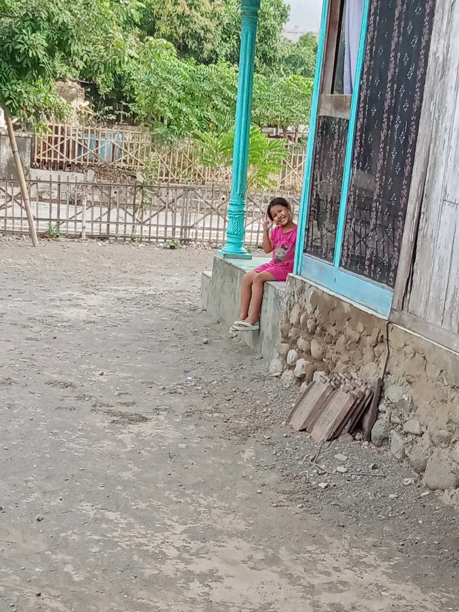 A young girl in a pink dress is sitting on a concrete ledge outside a rustic building. She is smiling and making a peace sign with her fingers. The building has a weathered wooden exterior and a blue-trimmed window. In the background, there is a bamboo fence and lush green trees. The ground is covered in dirt and small stones, typical of the environment in Selowogo, Indonesia. A pile of roofing tiles leans against the wall of the building.